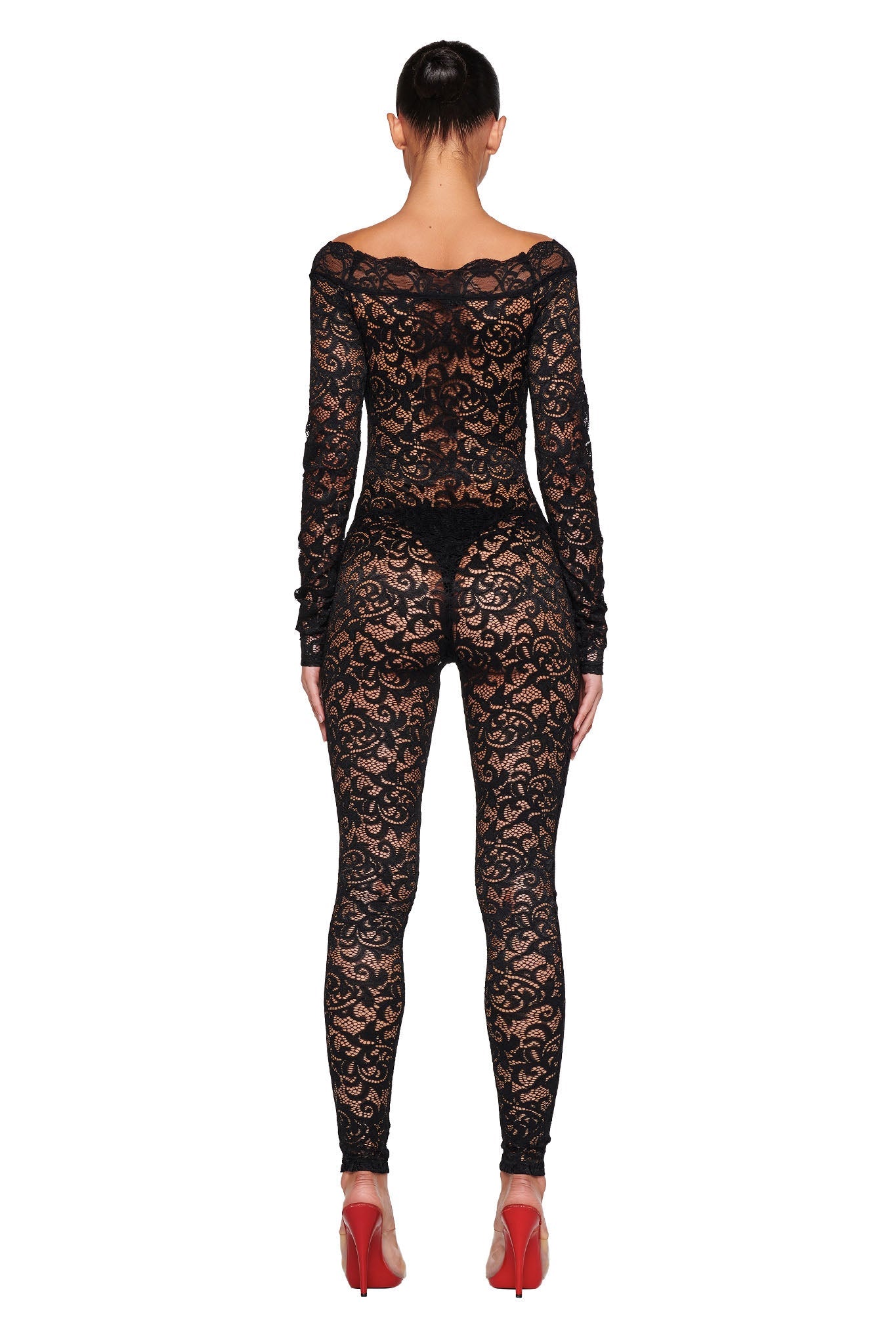 A person is standing with their back to the camera, wearing the long-sleeved, form-fitting GEMMA CATSUIT - BLACK from I.AM.GIA. The outfit features an intricate floral lace pattern. They are also adorned with red high-heeled shoes, and their hair is pulled back into a neat bun.