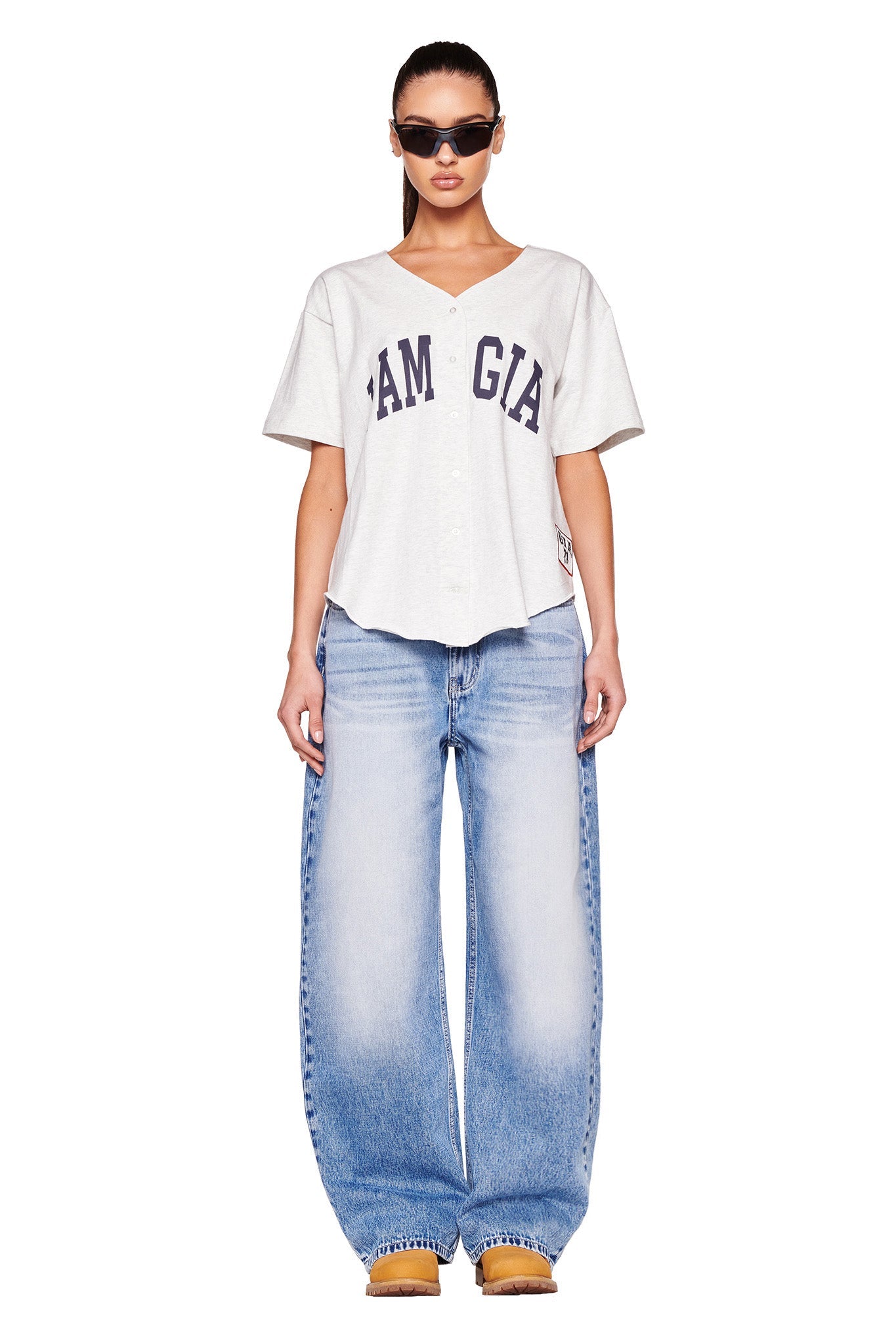 A person stands facing the camera wearing a grey LORRI JERSEY with "I.AM.GIA" printed on it, baggy light blue jeans, tan shoes, and black sunglasses. They have long hair pulled back and a neutral expression. The background is white.
