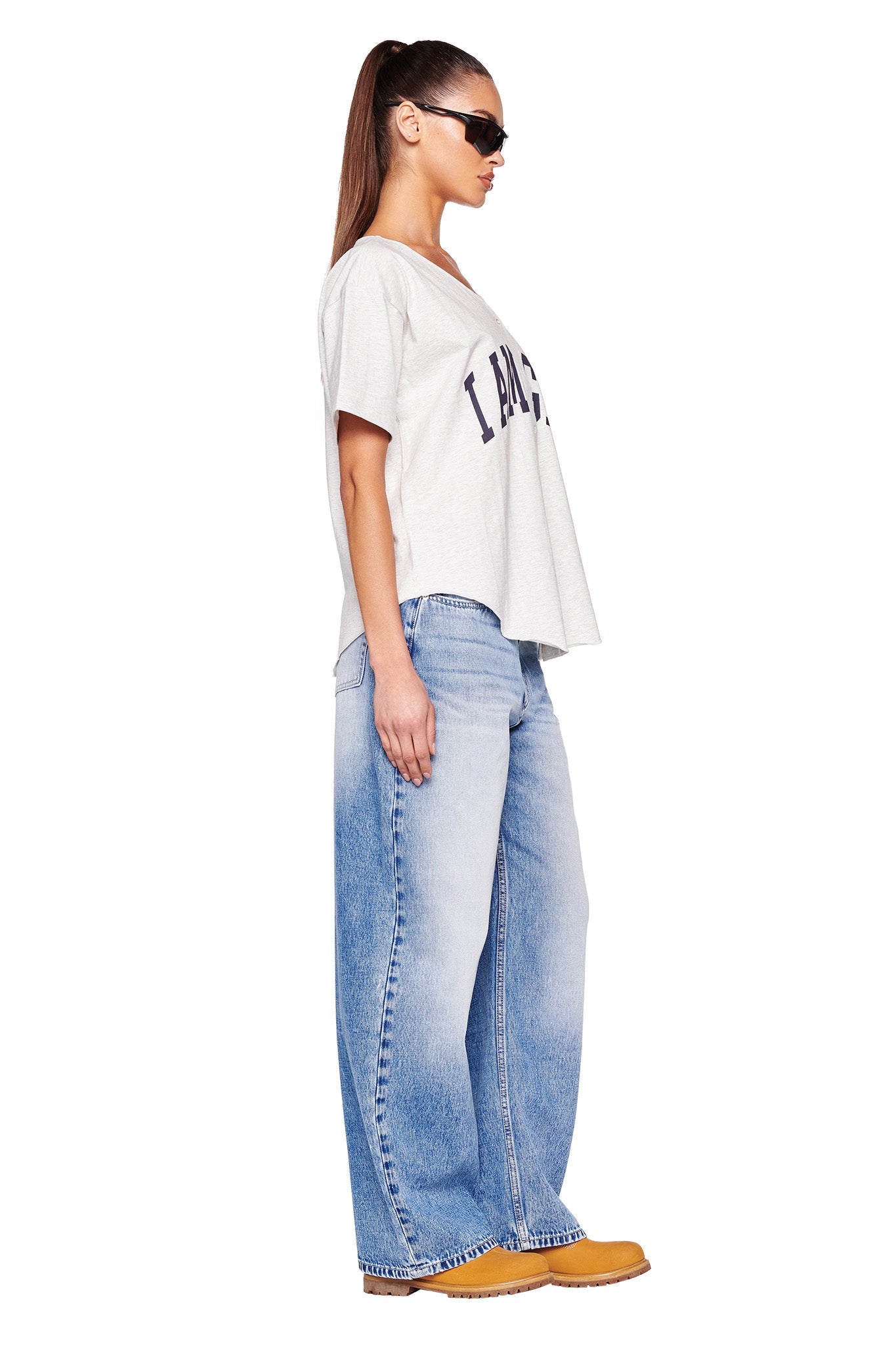 A woman stands in a side profile against a plain white background. She wears an I.AM.GIA loose-fitting LORRI Jersey in grey with text on the front, light blue wide-leg jeans, yellow platform shoes, and sunglasses. Her long hair is tied back in a ponytail.