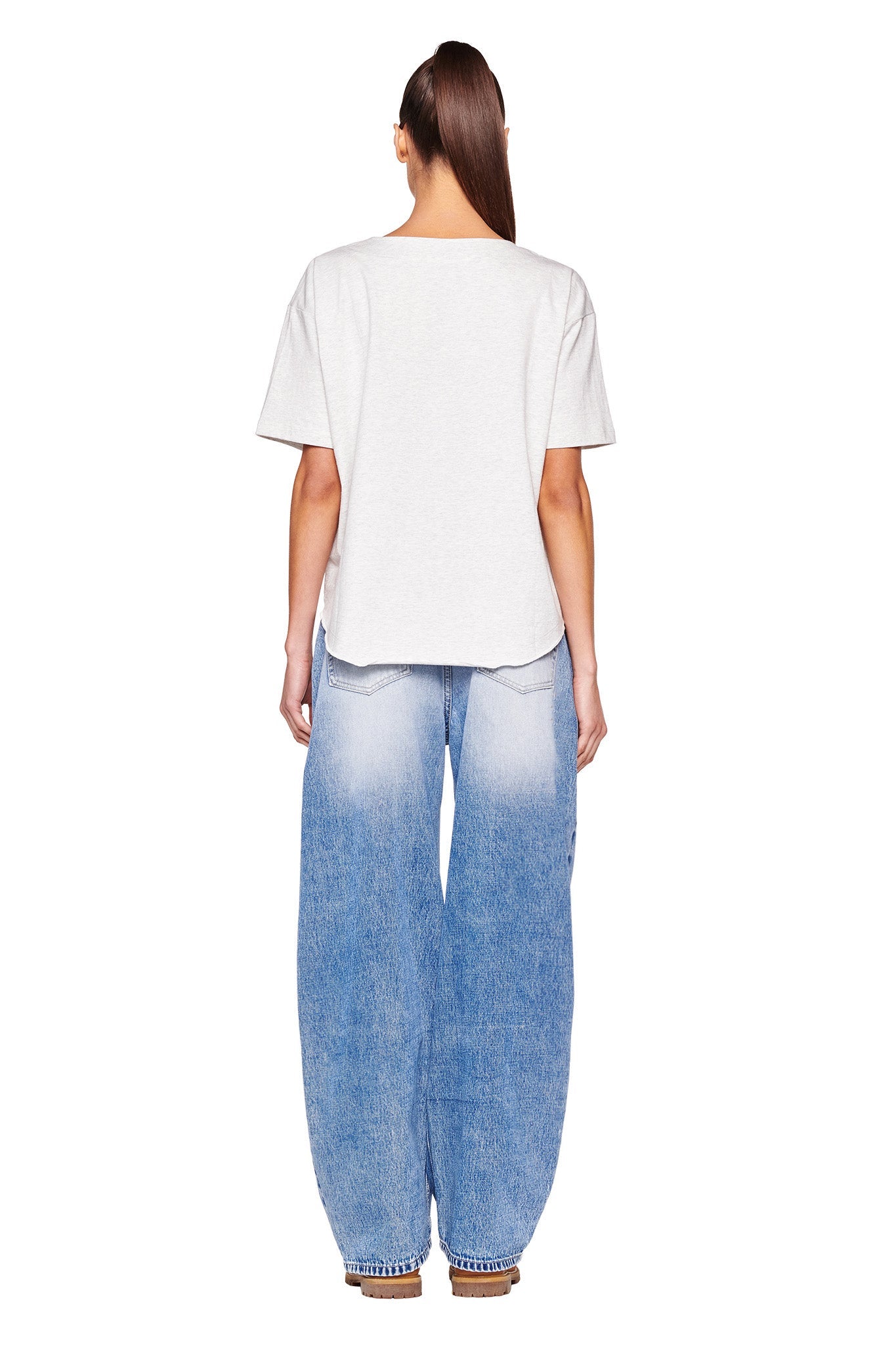 A person is standing with their back to the camera, long hair tied back in a ponytail. They are wearing a relaxed-fit white I.AM.GIA LORRI JERSEY - GREY T-shirt and baggy, light blue jeans that show a gradient effect towards the bottom. The person completes the look with brown sandals.