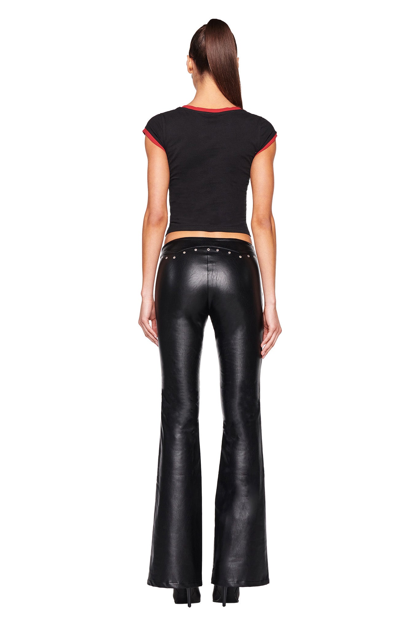 A person with long hair in a high ponytail is standing with their back to the camera. They are wearing the I.AM.GIA DORA TOP - BLACK, a short-sleeve crop top featuring red accents, paired with black, shiny, high-waisted flared pants adorned with stud details at the waistband. Black heels complete the edgy look.