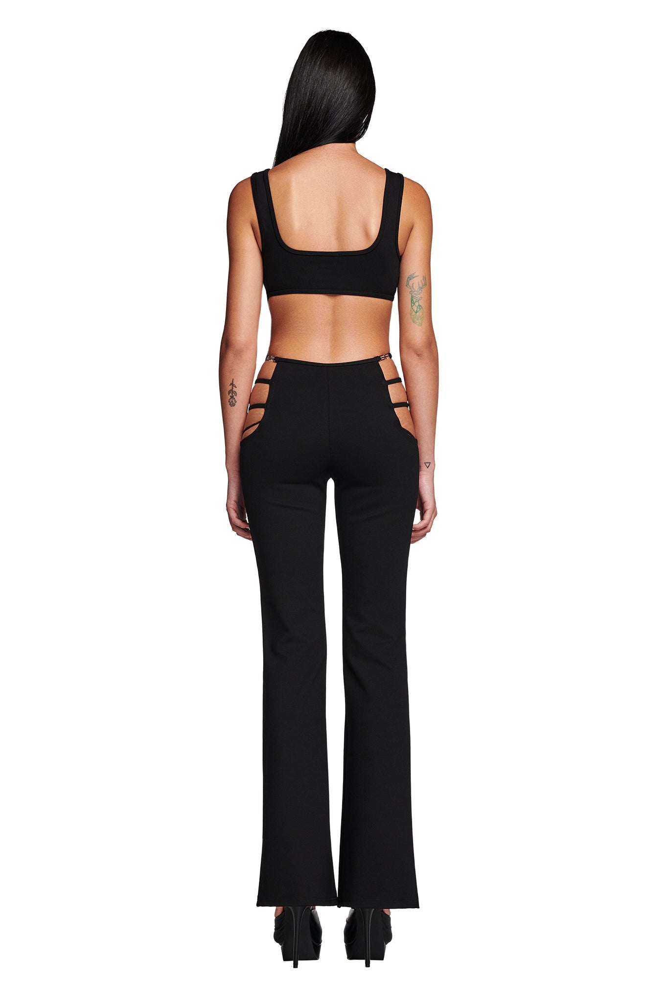 A woman stands facing away from the camera, showcasing her I.AM.GIA LUCID TOP - BLACK and high-waisted pants with multiple cutouts on the sides. She has long black hair and is wearing black platform shoes, flaunting tattoos on her upper left arm and left forearm.
