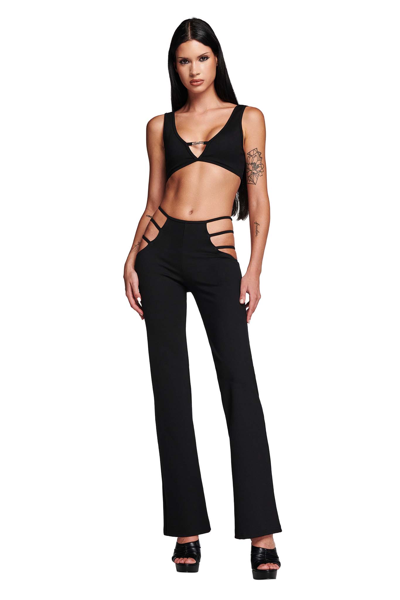 A woman with long dark hair is wearing the LUCID TOP - BLACK by I.AM.GIA, along with high-waisted black pants that feature hip cut-outs. She has a tattoo on her left upper arm and is standing against a plain white background, looking directly at the camera.