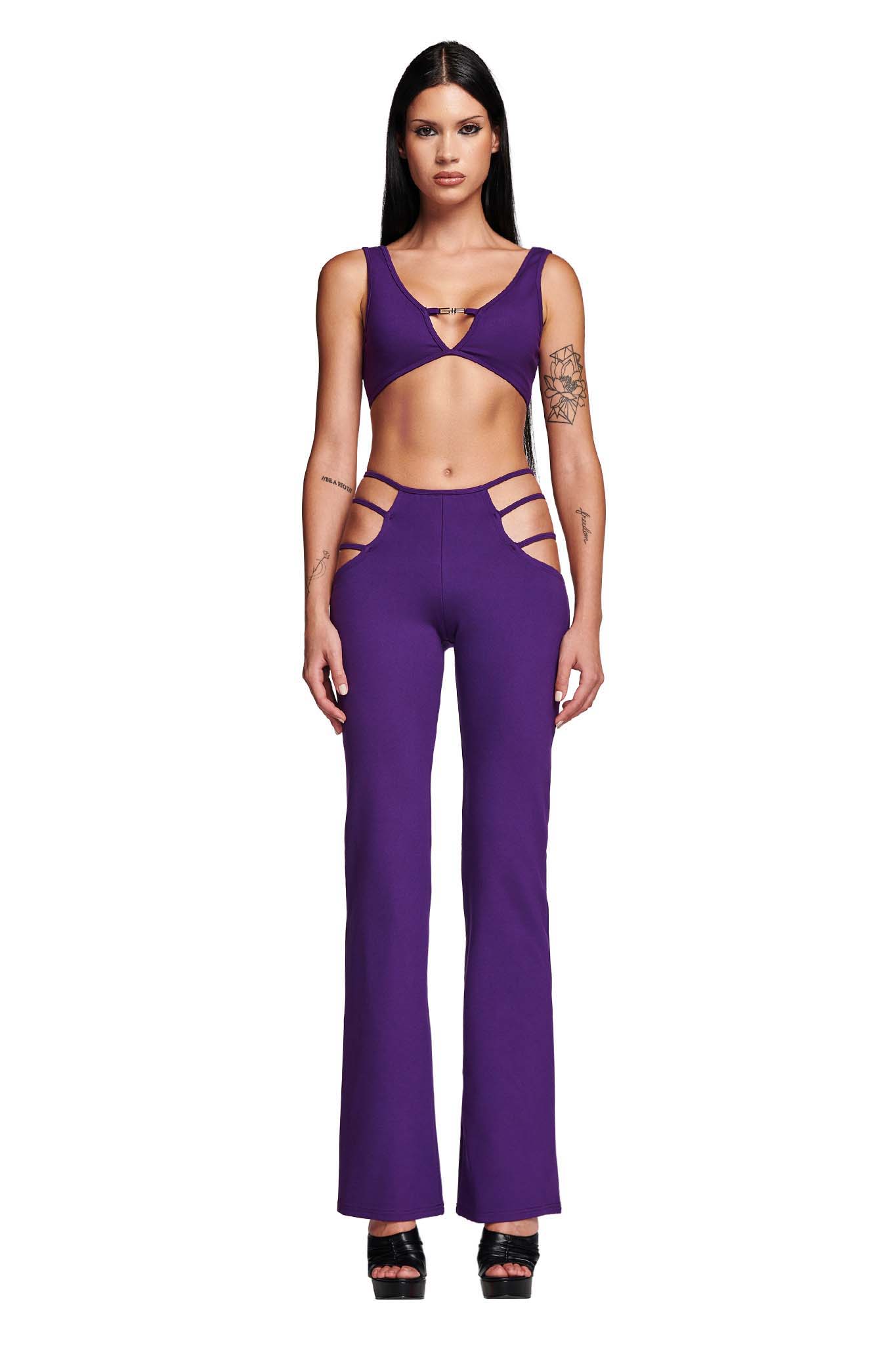 A woman is standing against a white background wearing the I.AM.GIA LUCID TOP in purple. The top has a deep V-neckline, and she pairs it with high-waisted pants featuring side cutouts on the hips. She is also wearing black high-heeled sandals. Her long, straight hair is styled neatly.