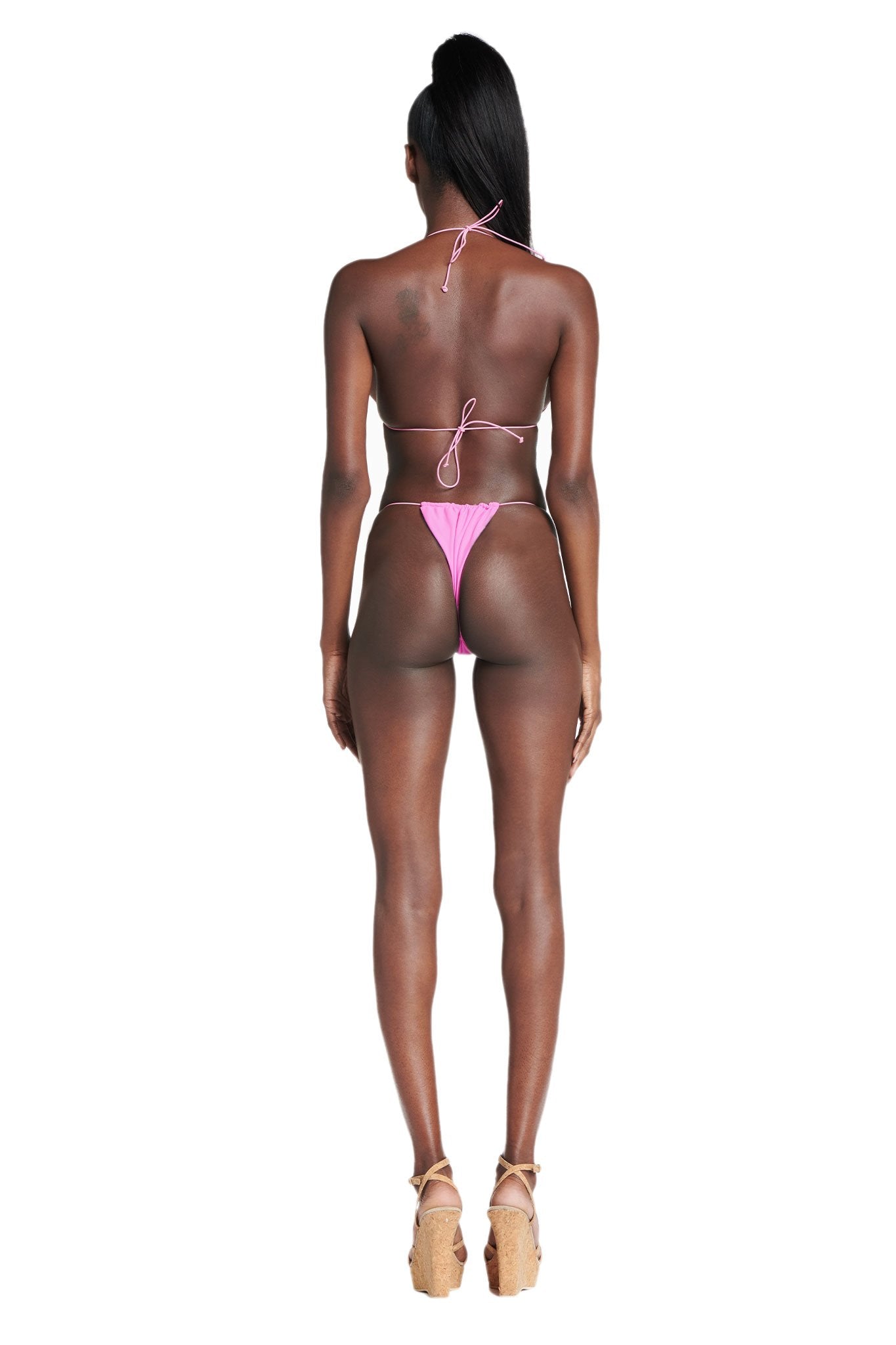 A woman with long dark hair faces away, wearing the I.AM.GIA BAMBI BIKINI BOTTOM in pink and wedge sandals against a plain white background.