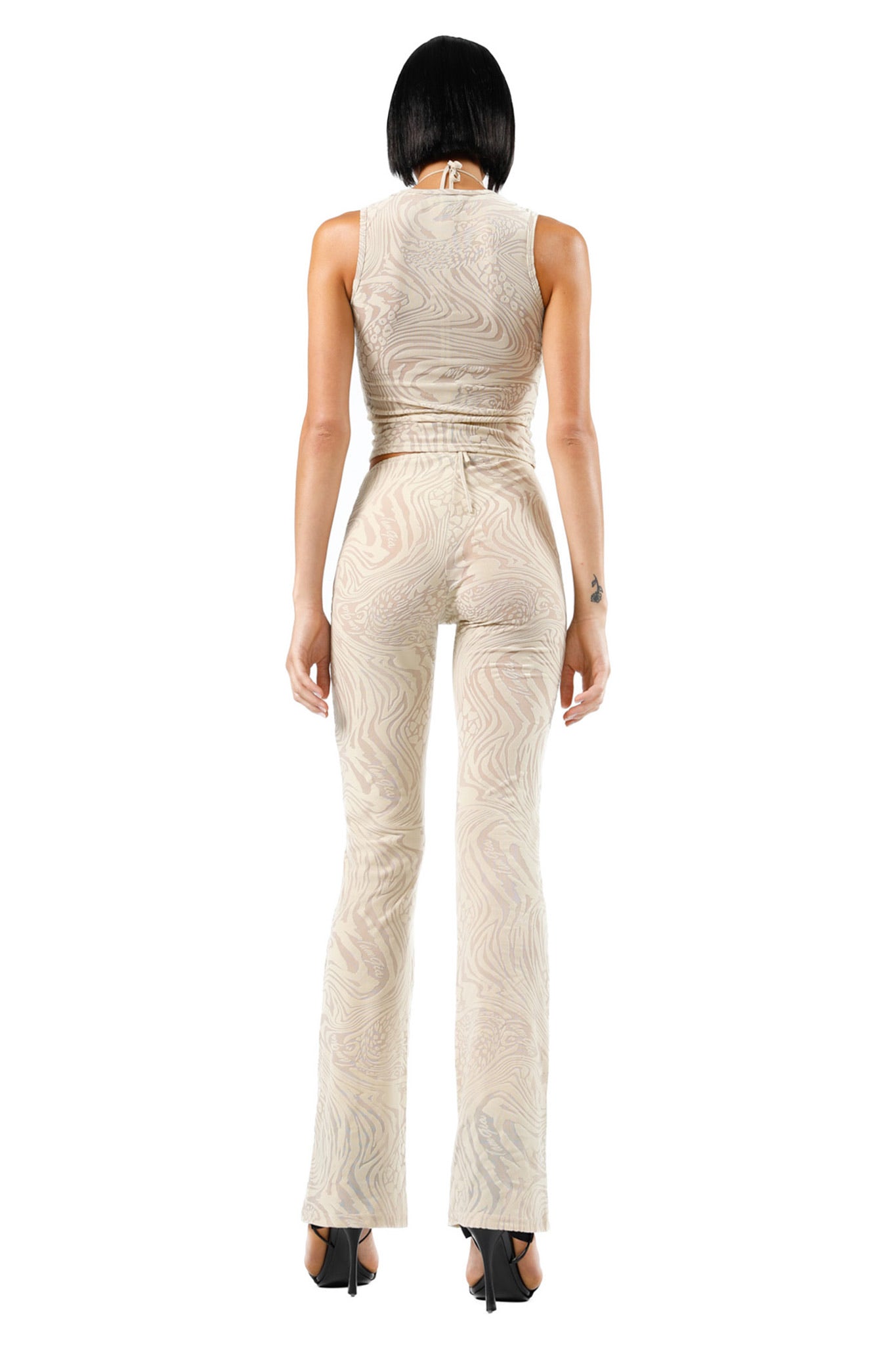 A woman with short, dark hair is facing away from the camera, dressed in a fitted and sleeveless I.AM.GIA's AMINA TOP in Tan Burnout with a subtle, swirling pattern. She pairs it with flared bottom pants and high-heeled shoes. A small tattoo adorns her left forearm.