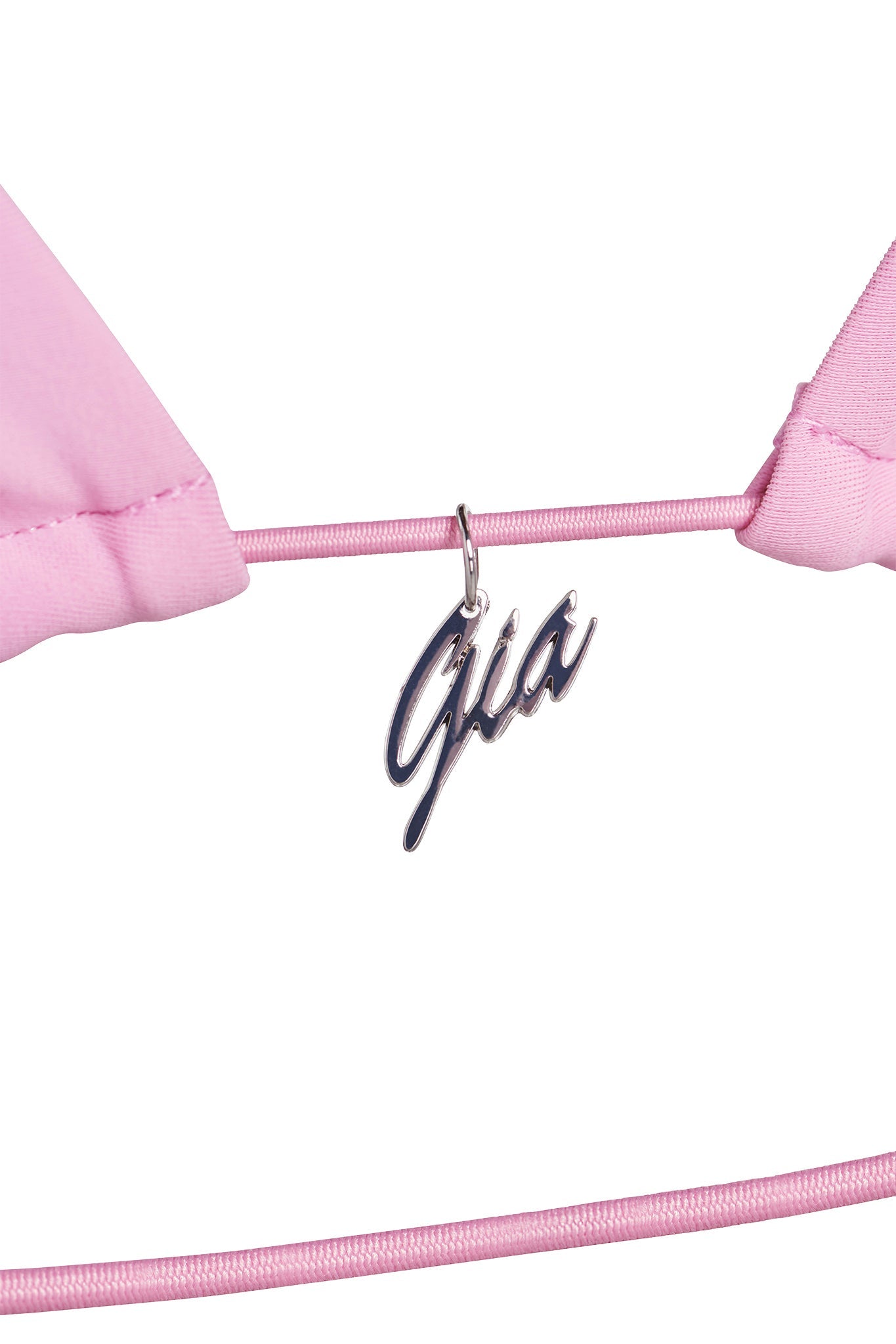 Close-up of a part of the BAMBI BIKINI TOP - PINK : BUBBLEGUM from I.AM.GIA, featuring a silver metal charm hanging from a thin pink elastic band. The charm is designed in a script font, forming the word "Ghia." The white background emphasizes the details of this I.AM.GIA piece.