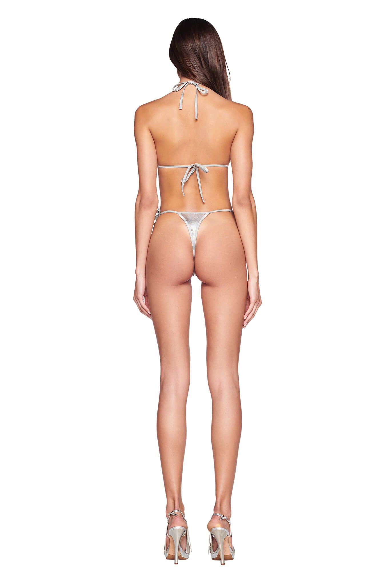 A woman is standing with her back to the camera, wearing the ALOHI BIKINI TOP - GREY : SILVER with tie straps at the neck and back from I.AM.GIA. She has long, straight hair and is wearing high-heeled sandals. The background is plain white.