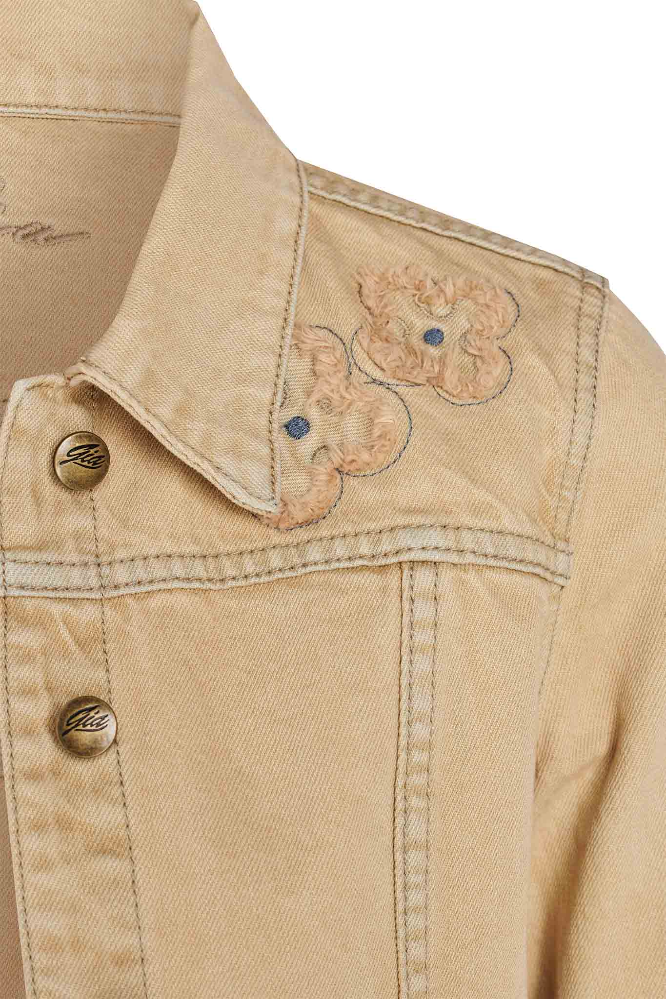 Close-up of the I.AM.GIA FAUNA JACKET - BEIGE, showcasing its light tan denim fabric, brass buttons, and floral embroidery on the left shoulder. The intricately detailed flowers have blue centers, adding a delicate touch of color. The jacket features a turned-down collar.