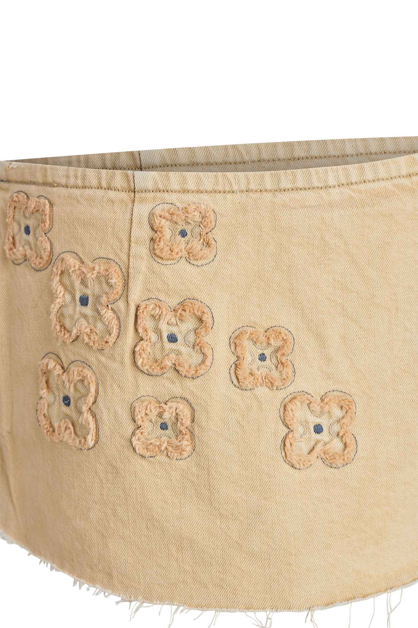 A close-up view reveals the beige fabric of the FAUNA MINI SKORT - BEIGE by I.AM.GIA, showcasing an unfinished edge adorned with light brown floral embroidery patterns. Delicately crafted details include embroidery stitches forming crosses at the centers of the flowers, embodying I.AM.GIA's distinct style.