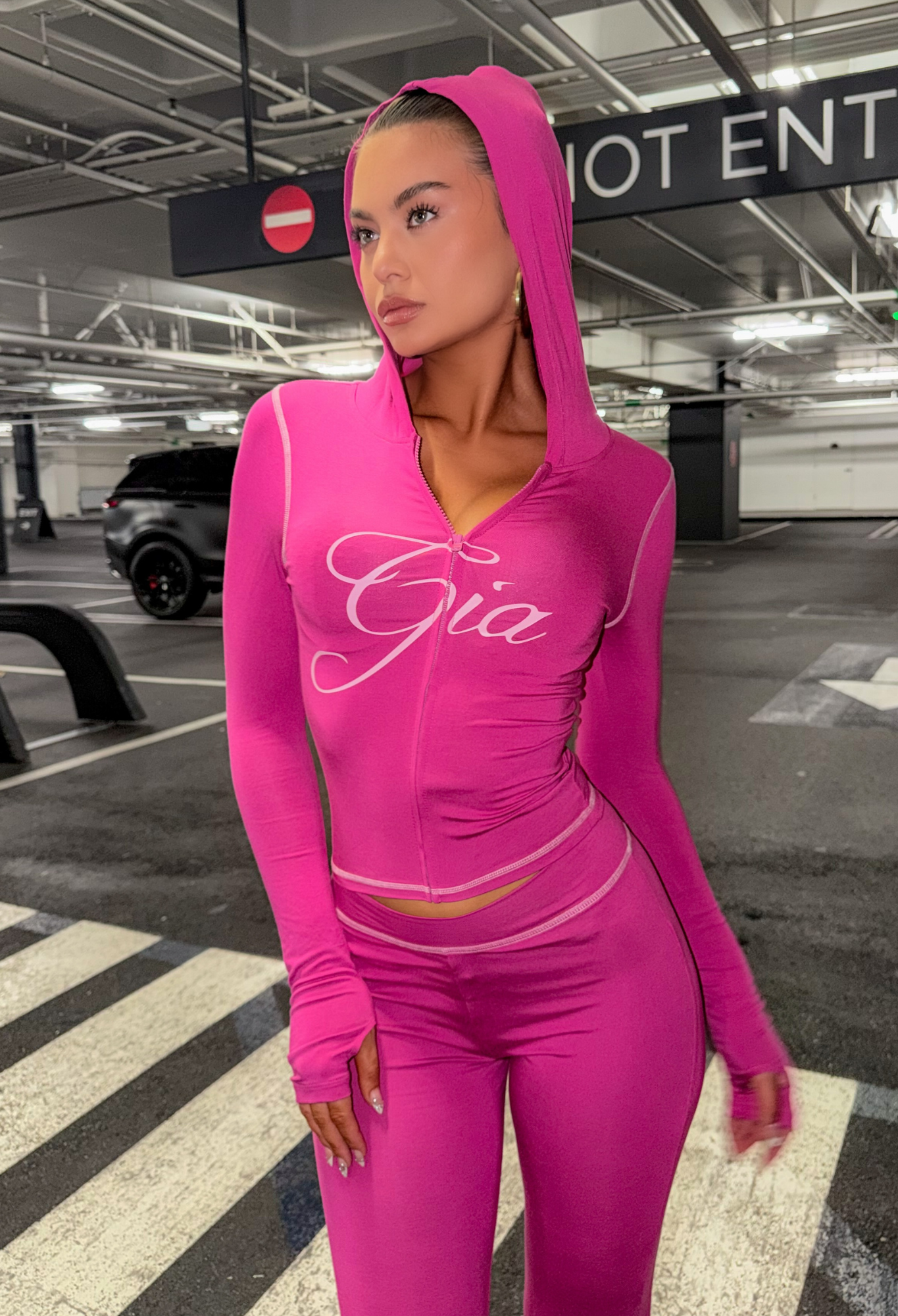 A woman wearing the I.AM.GIA BLARE HOODIE in raspberry stands on a crosswalk in a parking garage, looking to the side. The word "Gia" is written on her top.