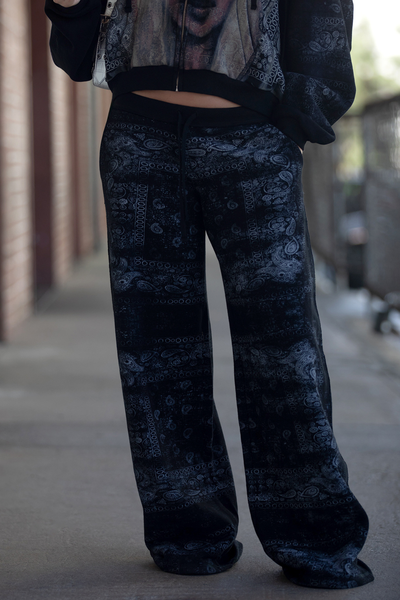 A person stands on a sidewalk showing off street style in the I.AM.GIA T-REX TRACKPANT - BLACK BANDANA and a coordinating jacket. The photo is cropped from the chest down, with brick and metal fencing visible in the background.