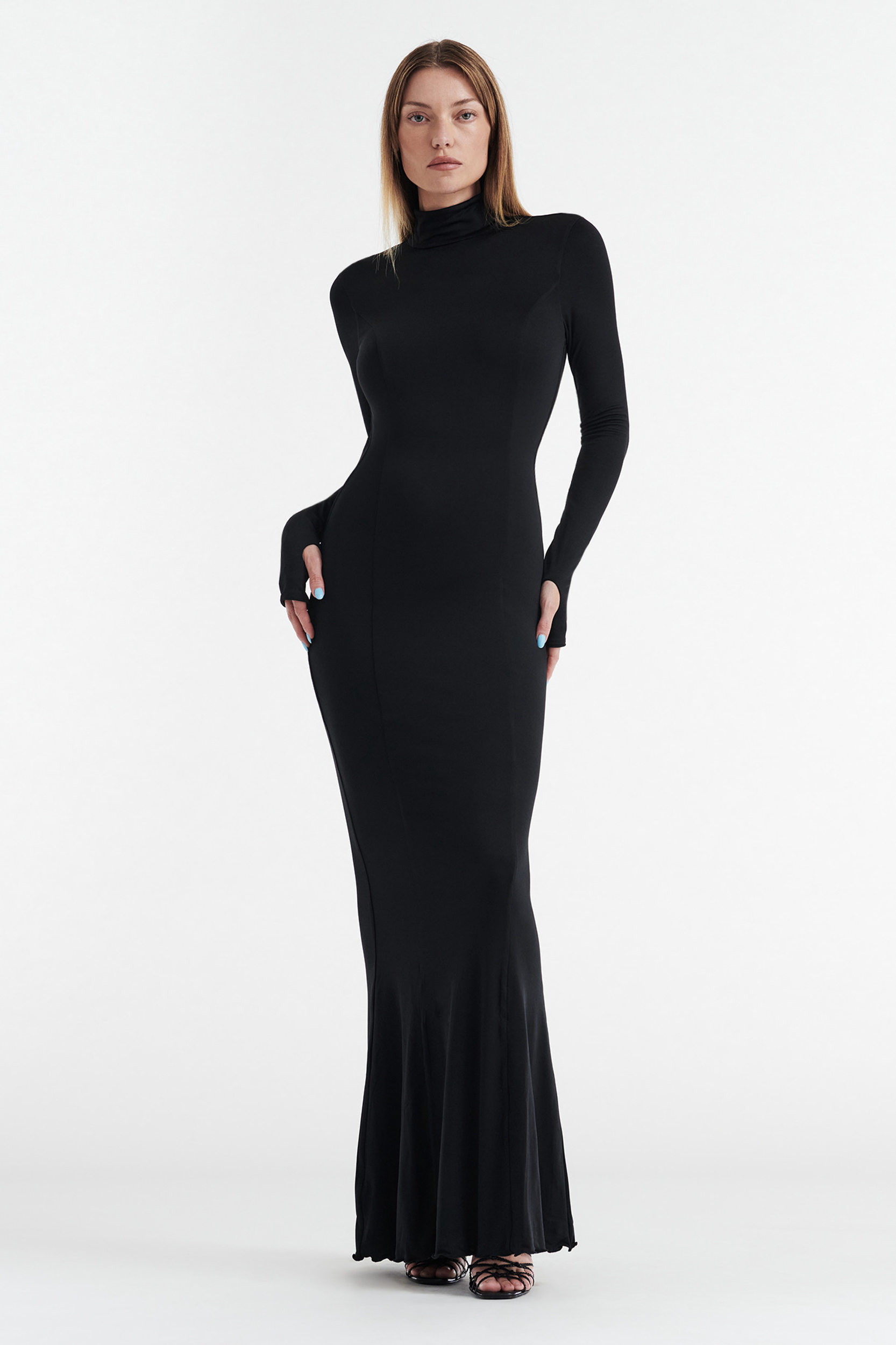A person stands facing forward in the CDP AUDREY Turtleneck Long Sleeve Maxi Dress in black, featuring a fitted silhouette and mermaid-style floor-length hem, with hands on hips against a plain white background.