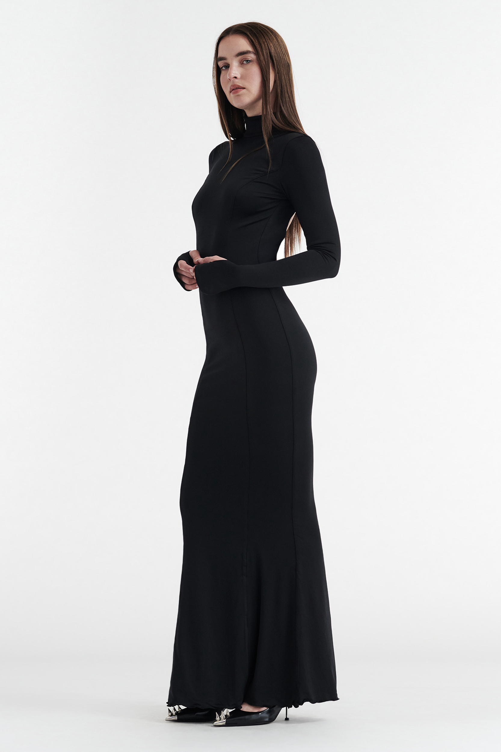 A woman with straight brown hair models the CDP AUDREY Turtleneck Long Sleeve Maxi Dress in black, featuring an open back, paired with black heels, and poses against a plain white background, facing slightly to the side.