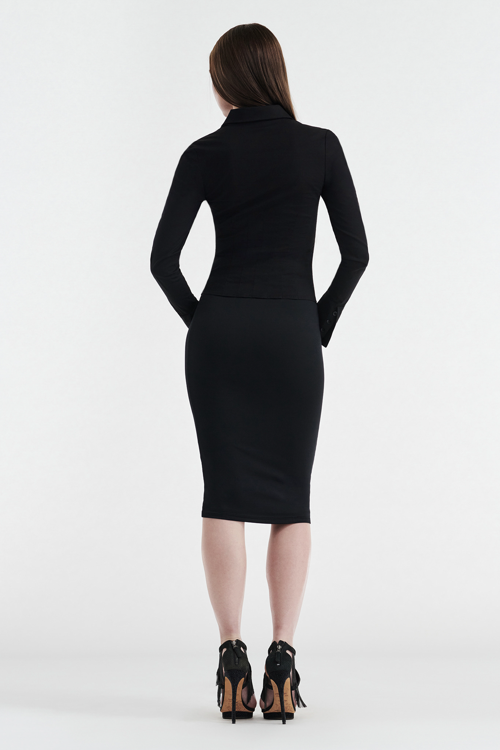 A woman with long brown hair faces away, wearing a fitted black long-sleeve top, CDP's GISELLE STRETCH MIDI TUBE SKIRT - BLACK, and black fringe-heeled shoes against a plain white background.