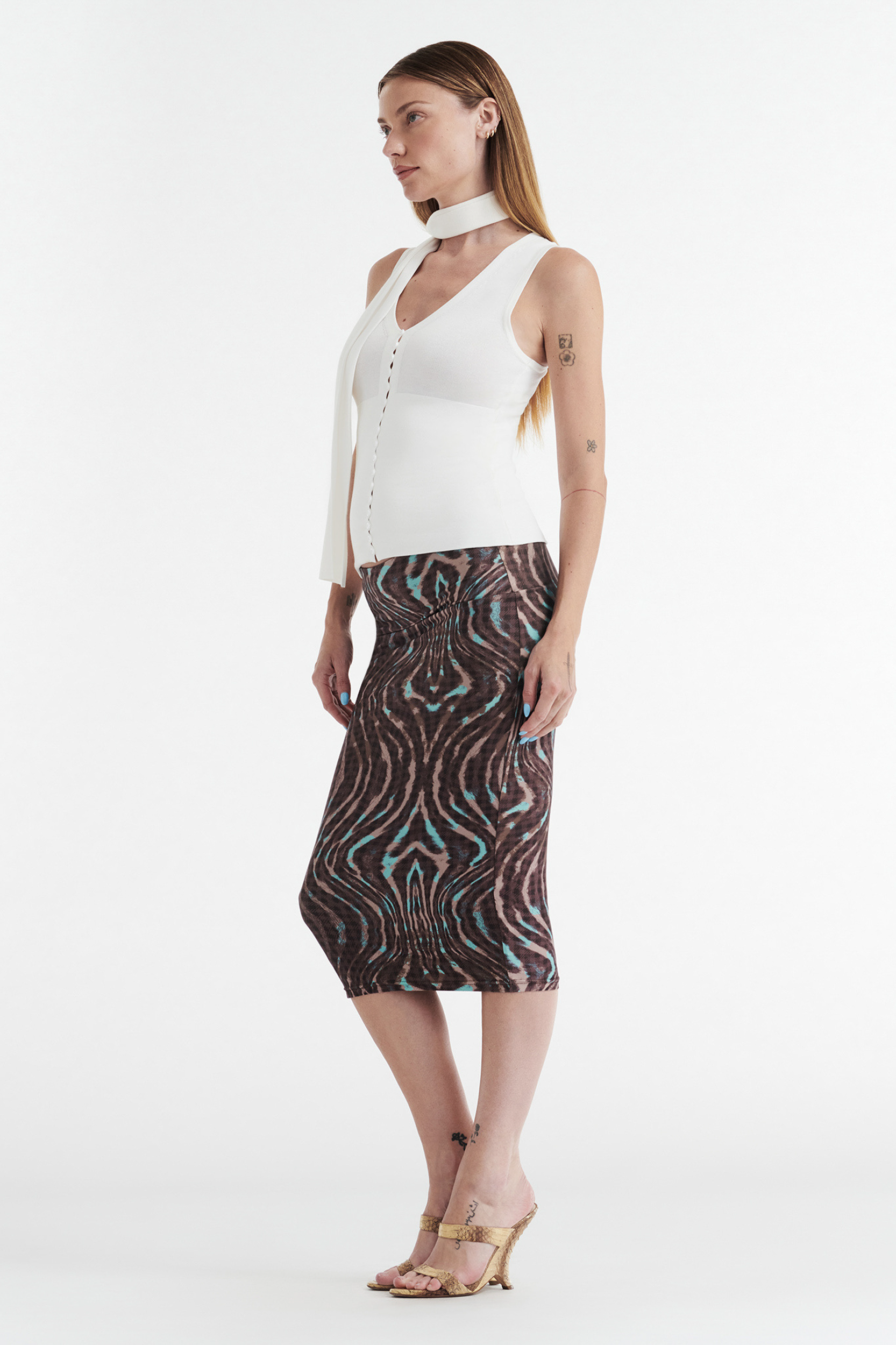 A woman with straight hair and tattoos models the CDP GISELLE STRETCH MIDI TUBE SKIRT - WARPED CHECK, paired with a sleeveless white top, neck scarf, and beige heeled sandals against a white background.