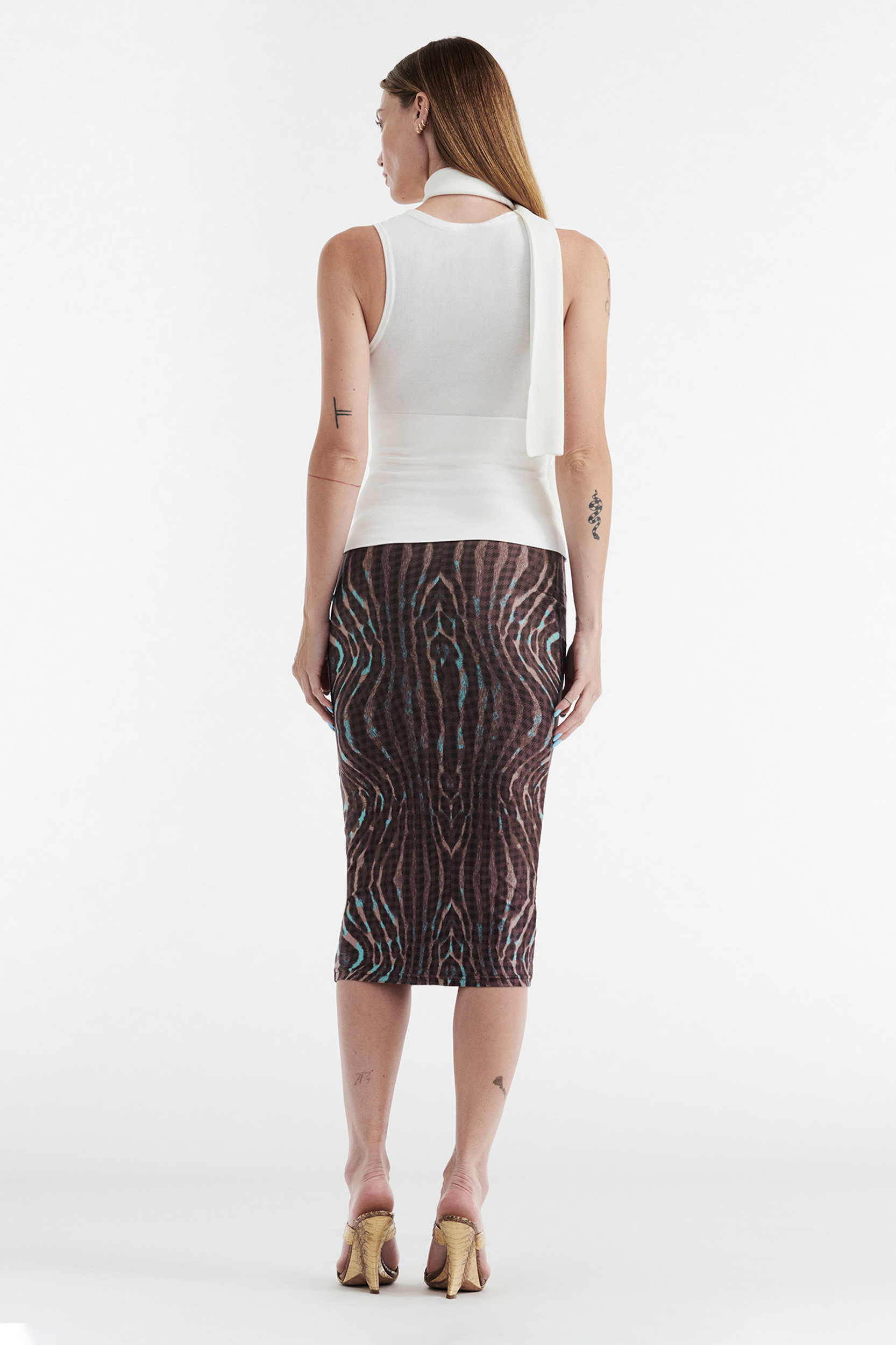 A woman with long, straight hair faces away from the camera, wearing a sleeveless white top and the CDP GISELLE Stretch Midi Tube Skirt in Warped Check, paired with heeled sandals.