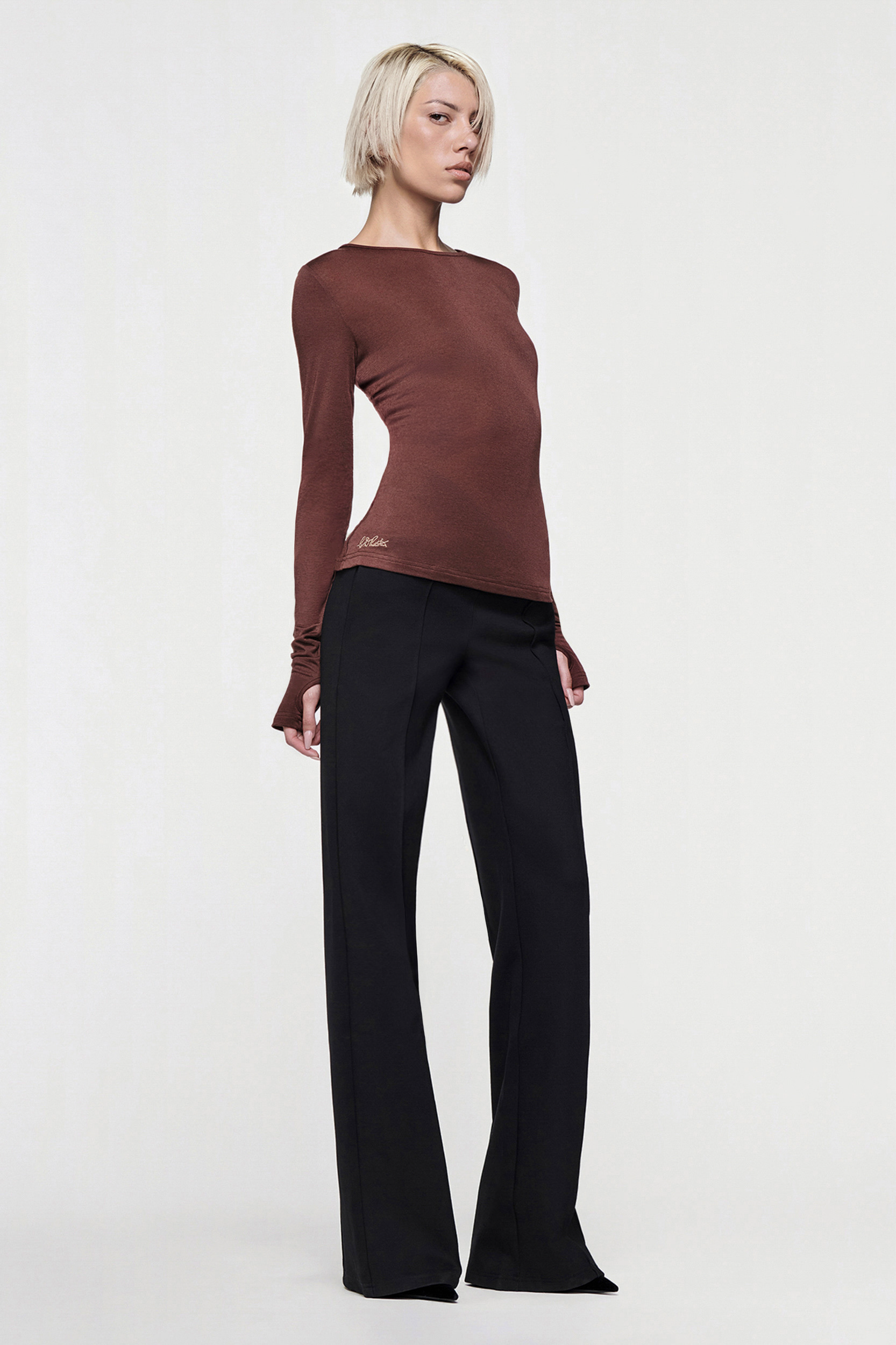 A person with short blonde hair wears the CDP CLAUDIA LONG SLEEVE BASIC TOP in chocolate and wide-leg black trousers, standing against a plain white background.