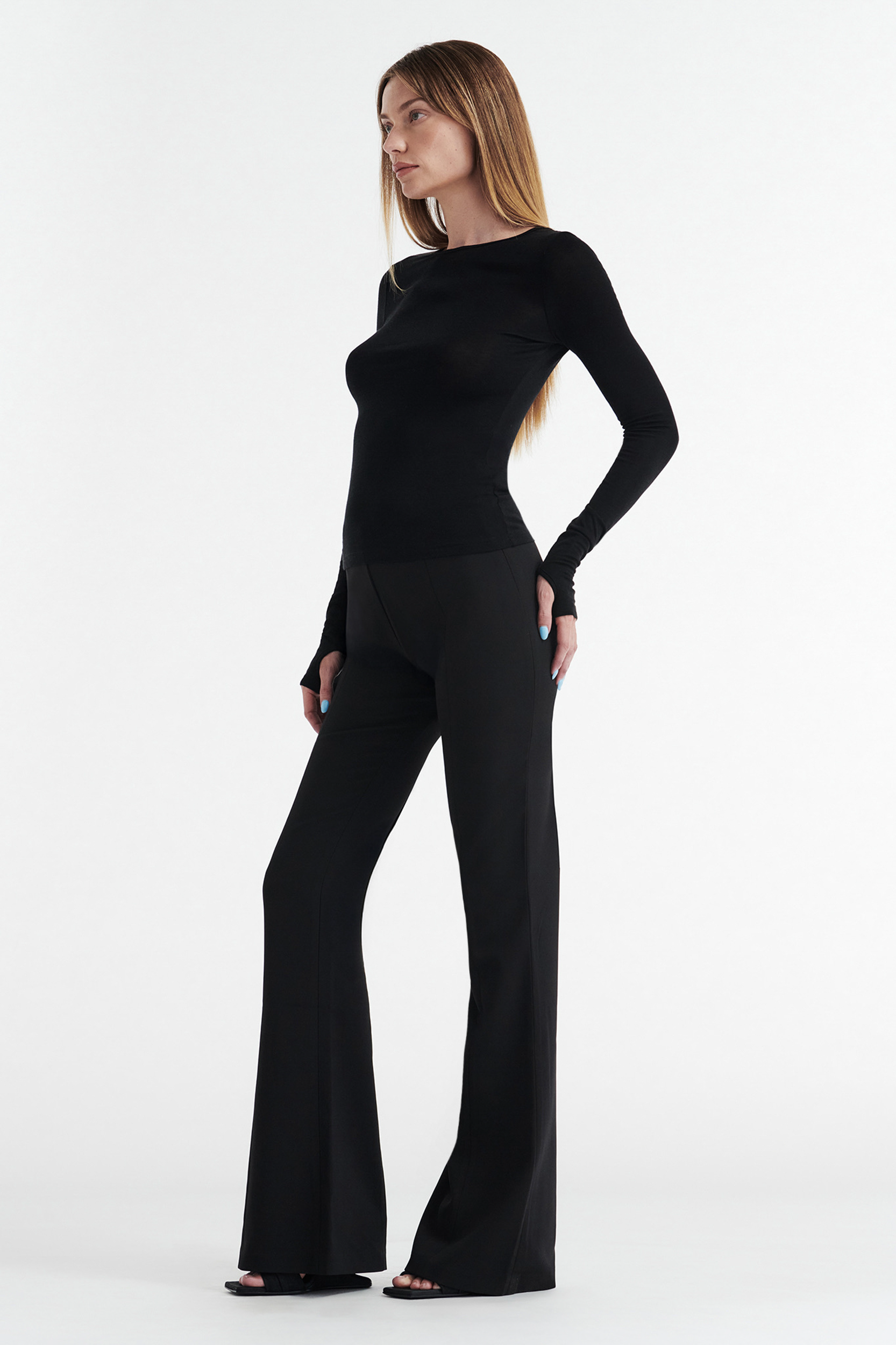 A woman with straight, light brown hair stands in profile against a white background, wearing the CDP CLAUDIA LONG SLEEVE BASIC TOP in black and black flared pants with open-toe shoes.