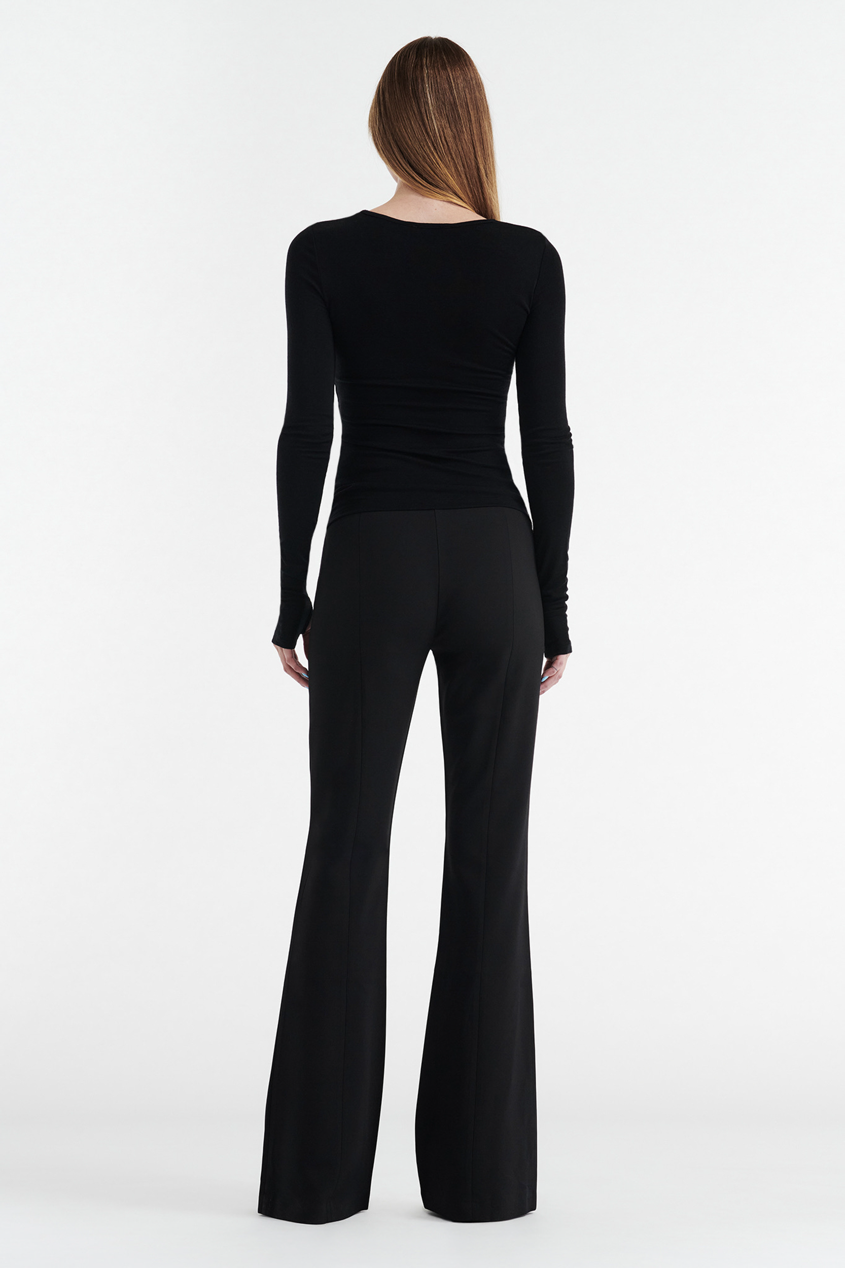 A woman with long straight hair faces away, wearing the CDP CLAUDIA LONG SLEEVE BASIC TOP - BLACK and black flared pants in a fitted silhouette, crafted from a lyocell wool blend, set against a plain white background.