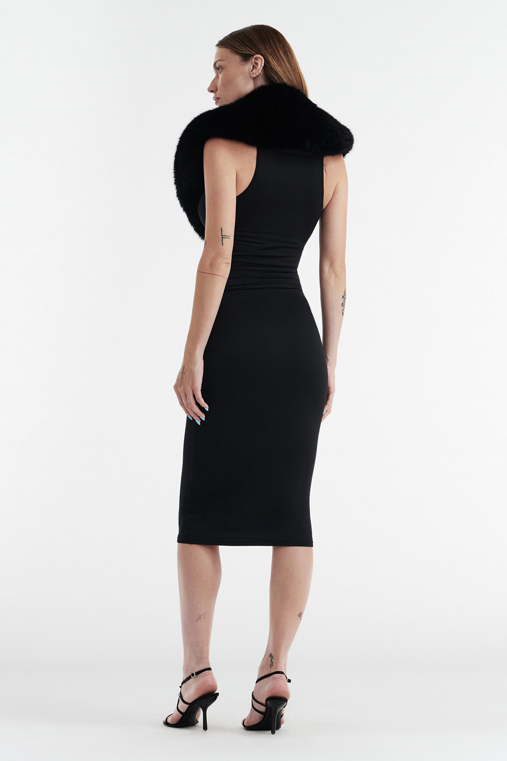 A woman stands facing away against a white background, wearing CDP’s SHIRLEY FAUX FUR COLLAR in black with a fitted sleeveless dress, black heeled sandals, and visible tattoos on her arms and ankles.