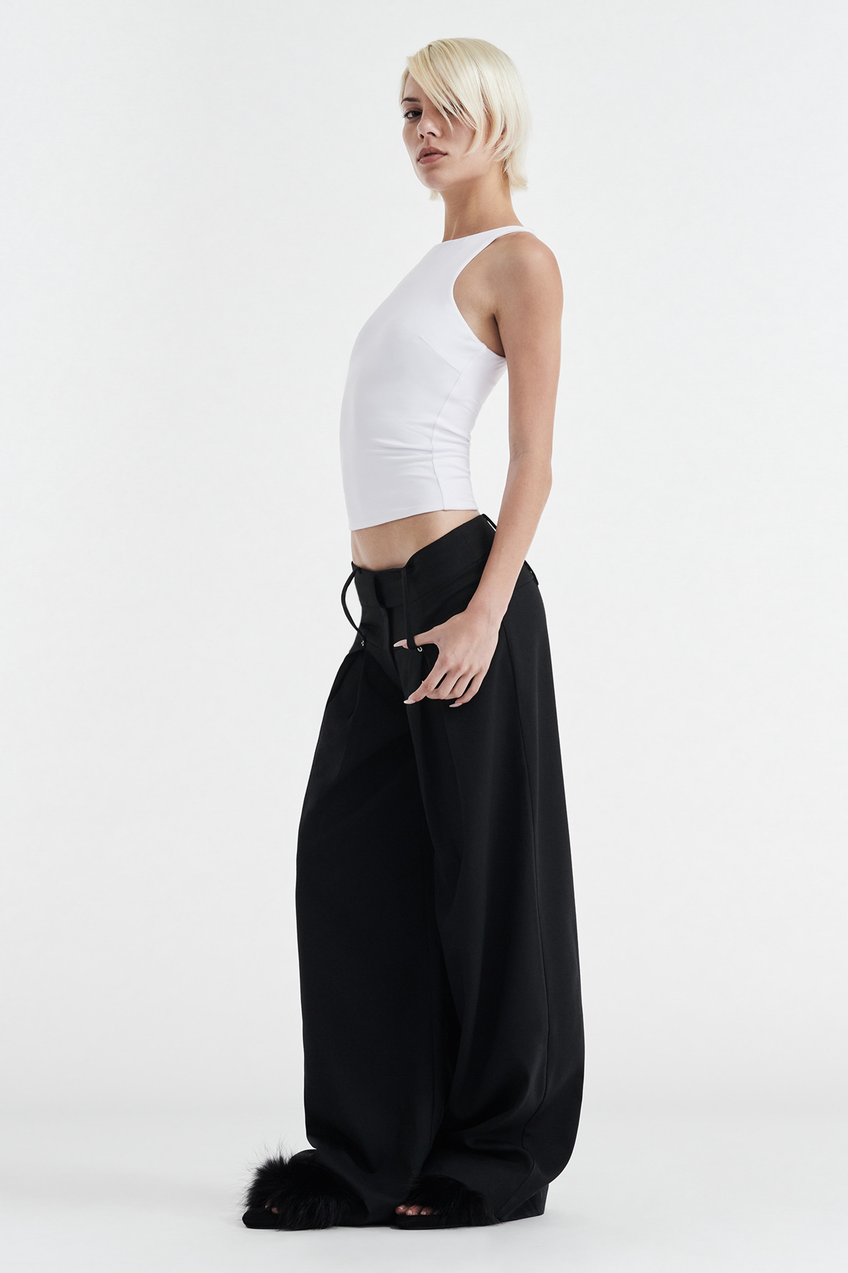 A person with short blond hair models the CDP CHARLOTTE WIDE LEG PLEAT PANT in black, paired with a fitted white sleeveless top and furry black shoes, standing sideways against a plain white background.