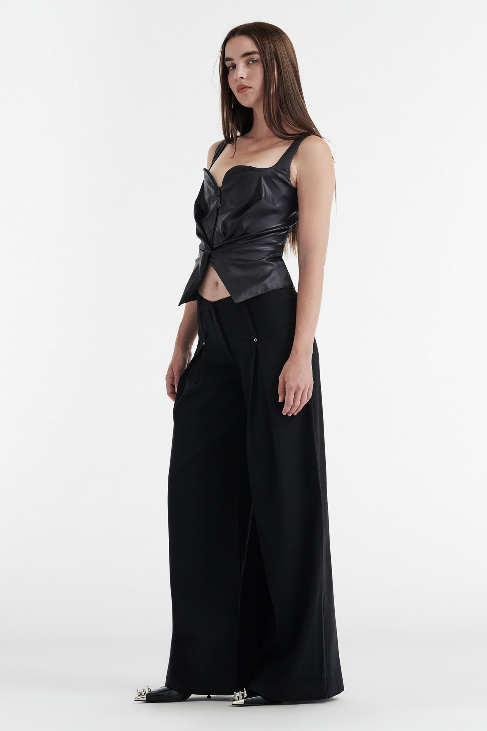 A woman with straight brown hair wears the CDP CHARLOTTE WIDE LEG PLEAT PANT in black, paired with a sculptural black leather strapless top and black pointed shoes, standing against a plain white background.