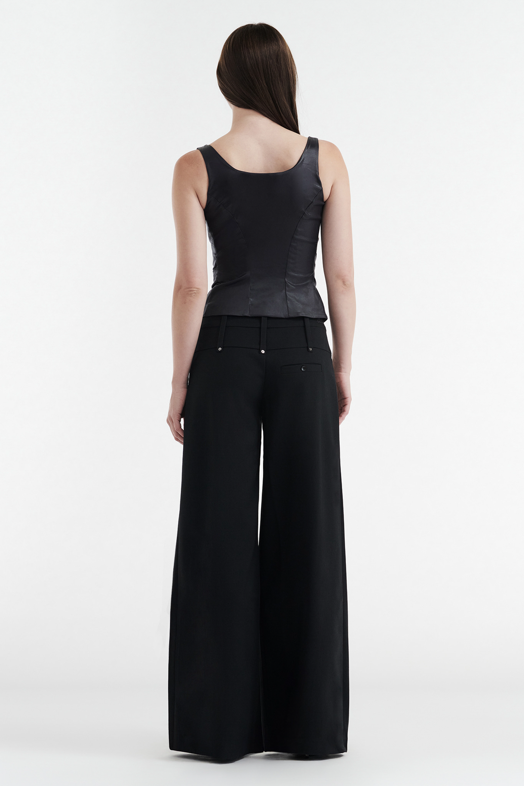 A woman faces away in a sleeveless black top and CDP’s CHARLOTTE WIDE LEG PLEAT PANT - BLACK. Her straight brown hair falls past her shoulders, set against a plain white background.