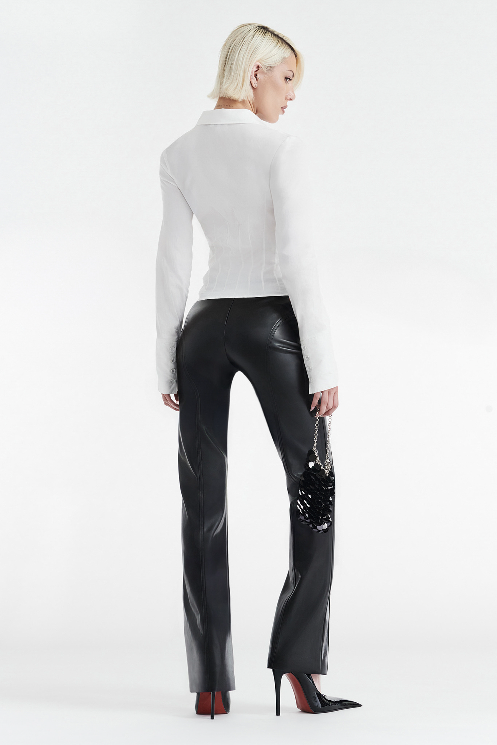 A person with short blonde hair faces away, wearing the CDP ZOE LONG SLEEVE COTTON BUTTON UP SHIRT in white, black leather pants, black high heels, and holding a black handbag against a plain white background.