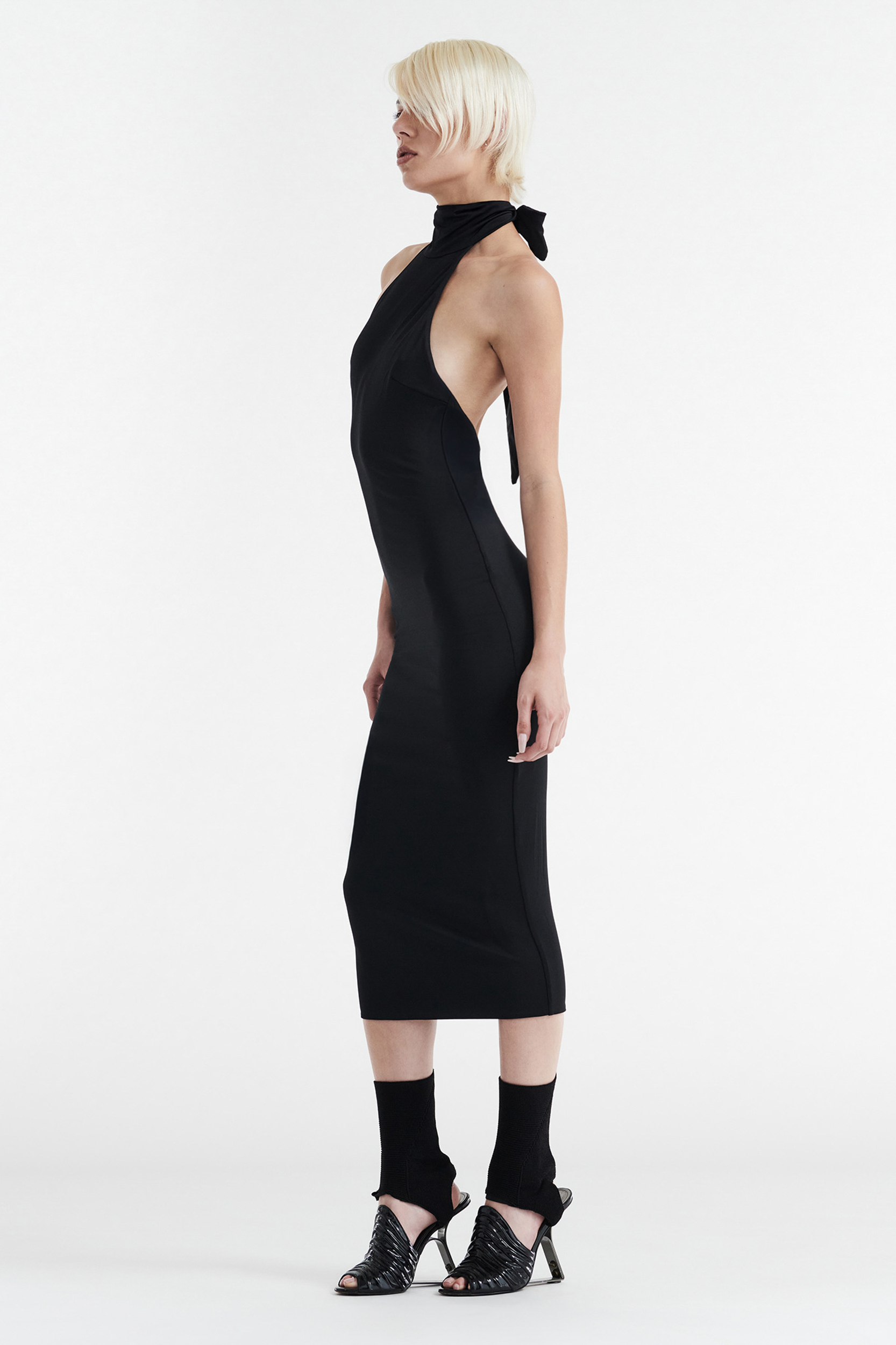 A person with short platinum blonde hair wears the CDP CLEO HALTER TIE MIDI DRESS in black, paired with black socks and open-toe heeled sandals, standing in profile against a plain white background.