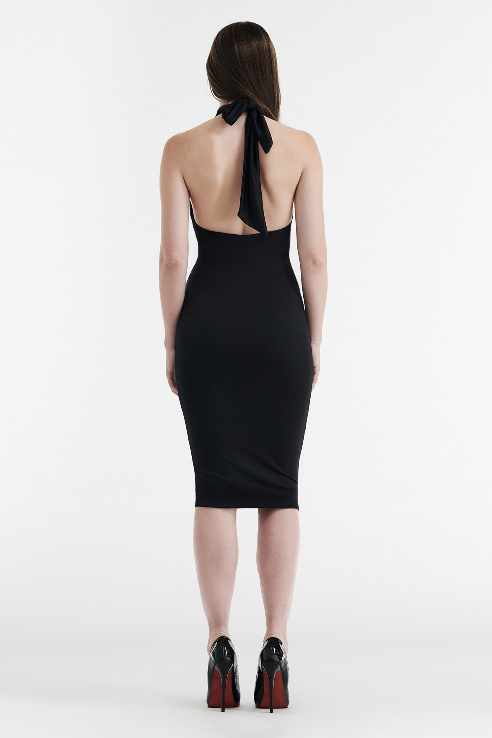 A woman with straight brown hair wears the CDP CLEO HALTER TIE MIDI DRESS in black, featuring a fitted bodycon silhouette, open back, and large halter turtleneck bow. She pairs it with black heels against a plain white background.