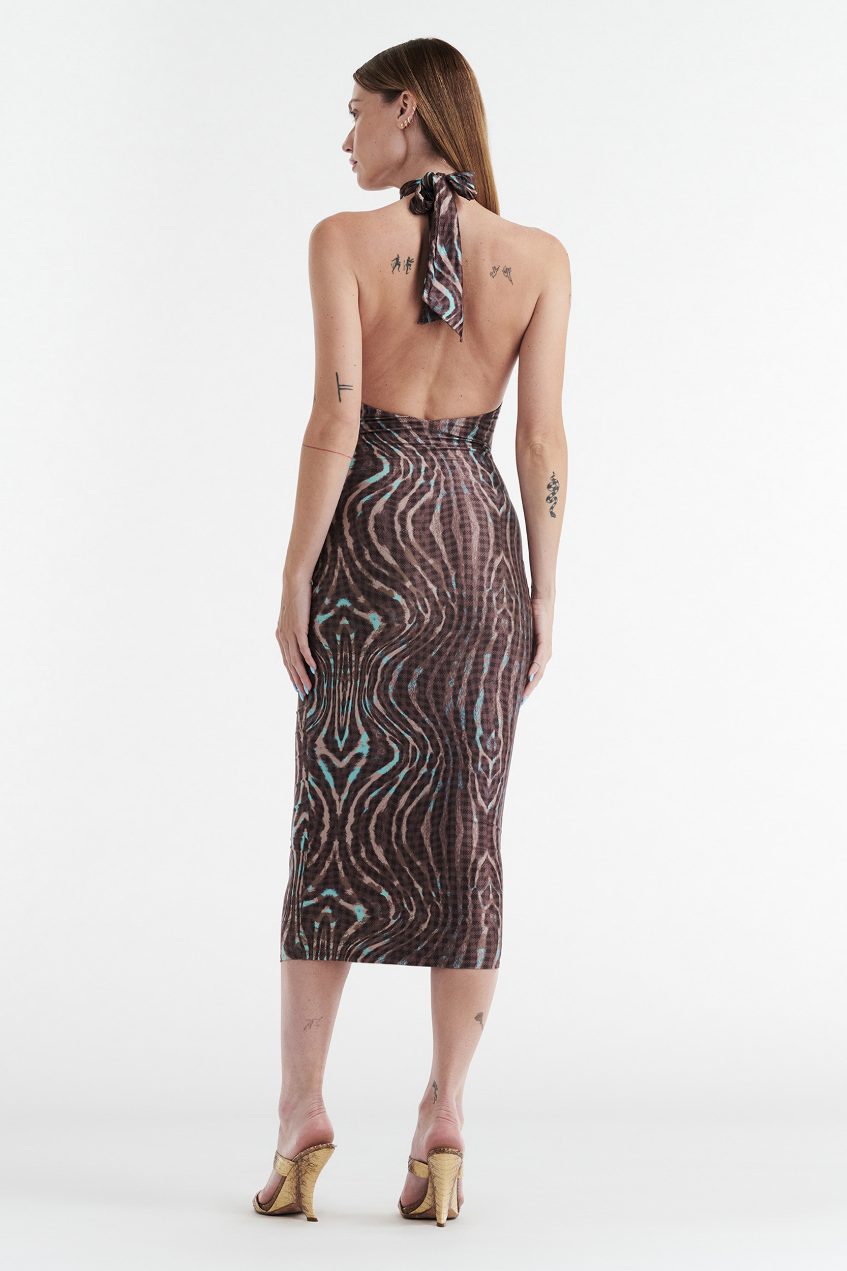 A woman with tattoos wears the CDP CLEO HALTER TIE MIDI DRESS - WARPED CHECK, featuring a brown and teal abstract print, halter turtleneck, open back, and mid-calf length. She pairs it with heeled sandals.