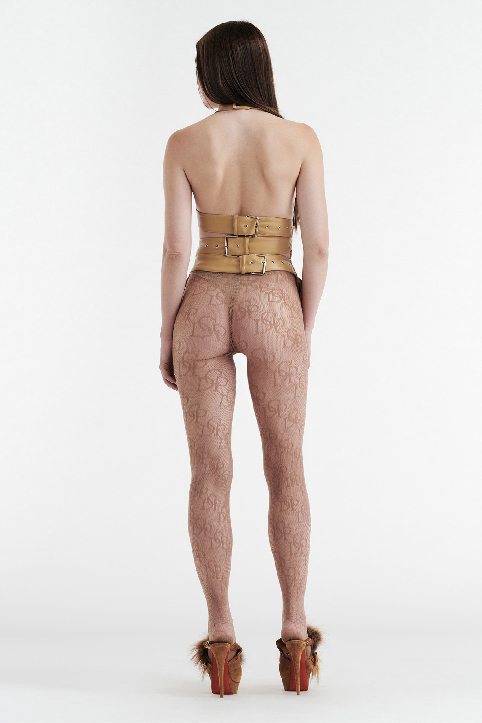A woman with long brown hair wears the CDP INES Asymmetrical Faux Leather Top in tan, styled with sheer patterned leggings and fur-accented high heels, standing against a plain white background.
