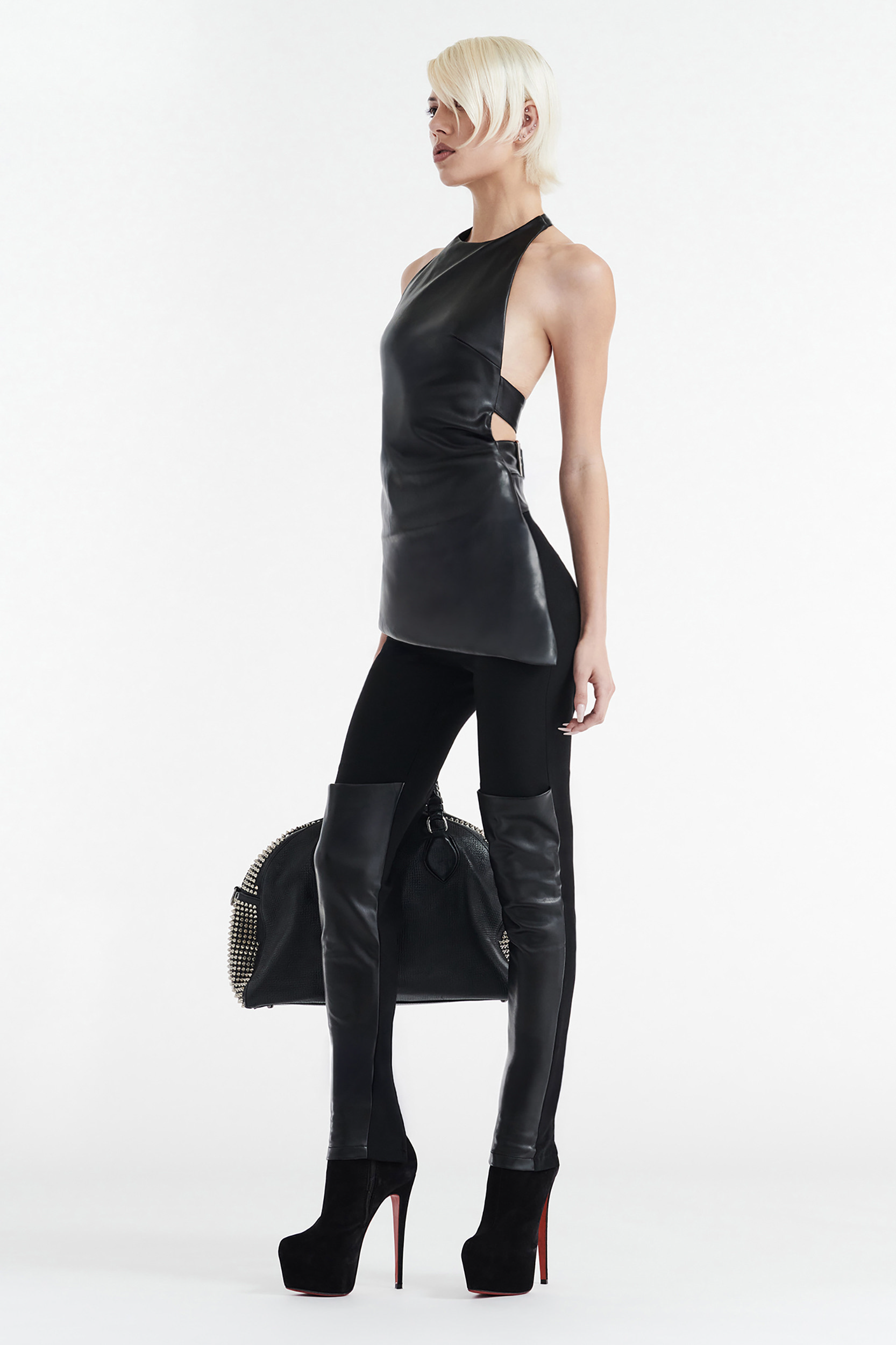 A person with short platinum blonde hair wears the CDP INES ASYMMETRICAL FAUX LEATHER TOP in black, black leggings with leather thigh-high panels, black high-heeled platform shoes, and holds a large black handbag against a white background.