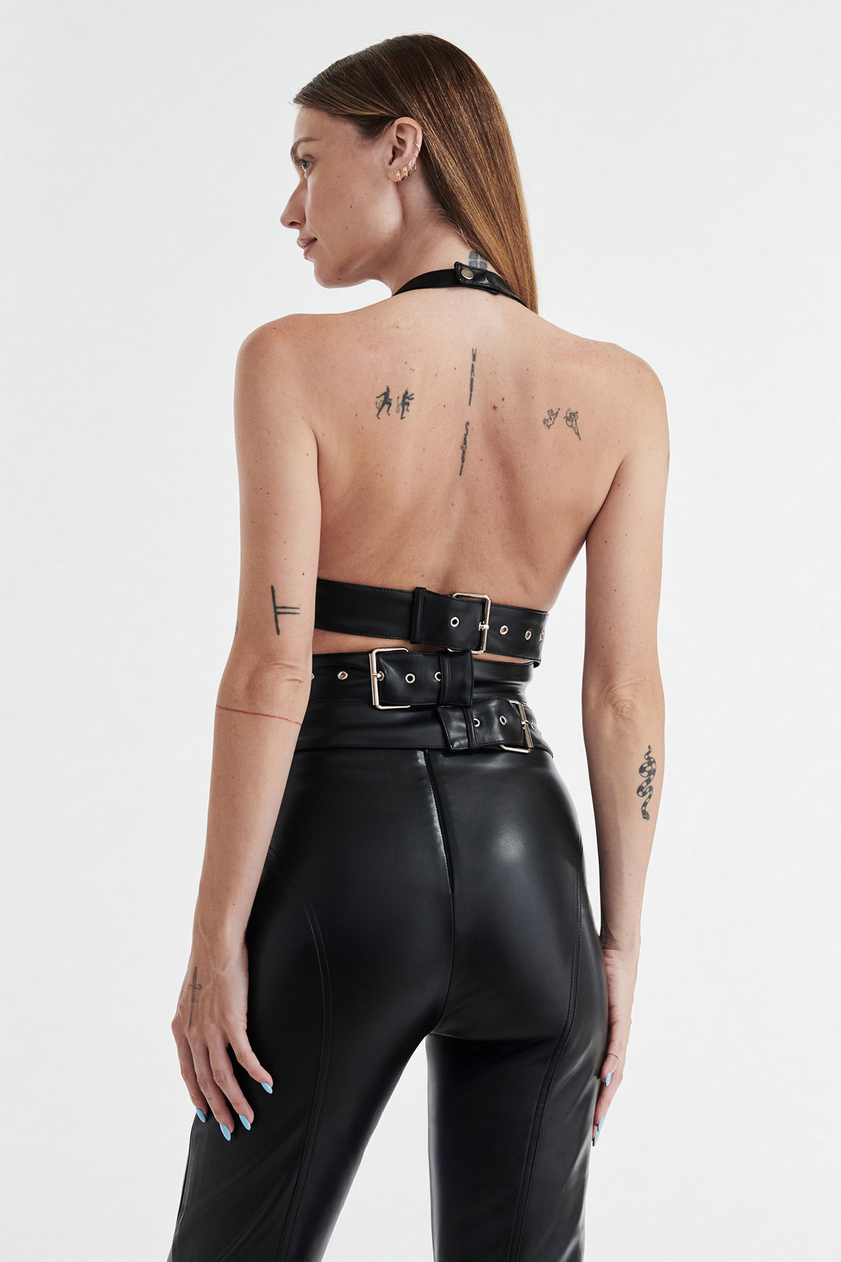 A woman with straight brown hair and visible tattoos is shown from the back wearing the CDP INES Asymmetrical Faux Leather Top in black, paired with high-waisted black leather pants, against a plain white background.