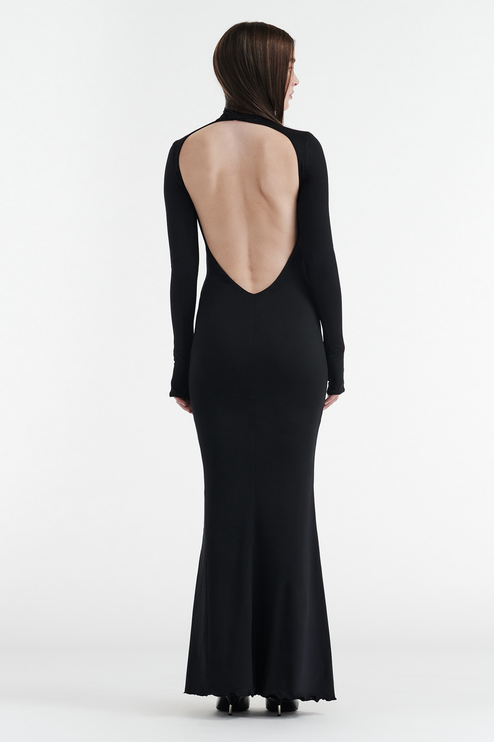 A person faces away in the CDP AUDREY TURTLENECK LONG SLEEVE MAXI DRESS - BLACK, featuring a dramatic open back and sleek mermaid-style hem against a plain white background.