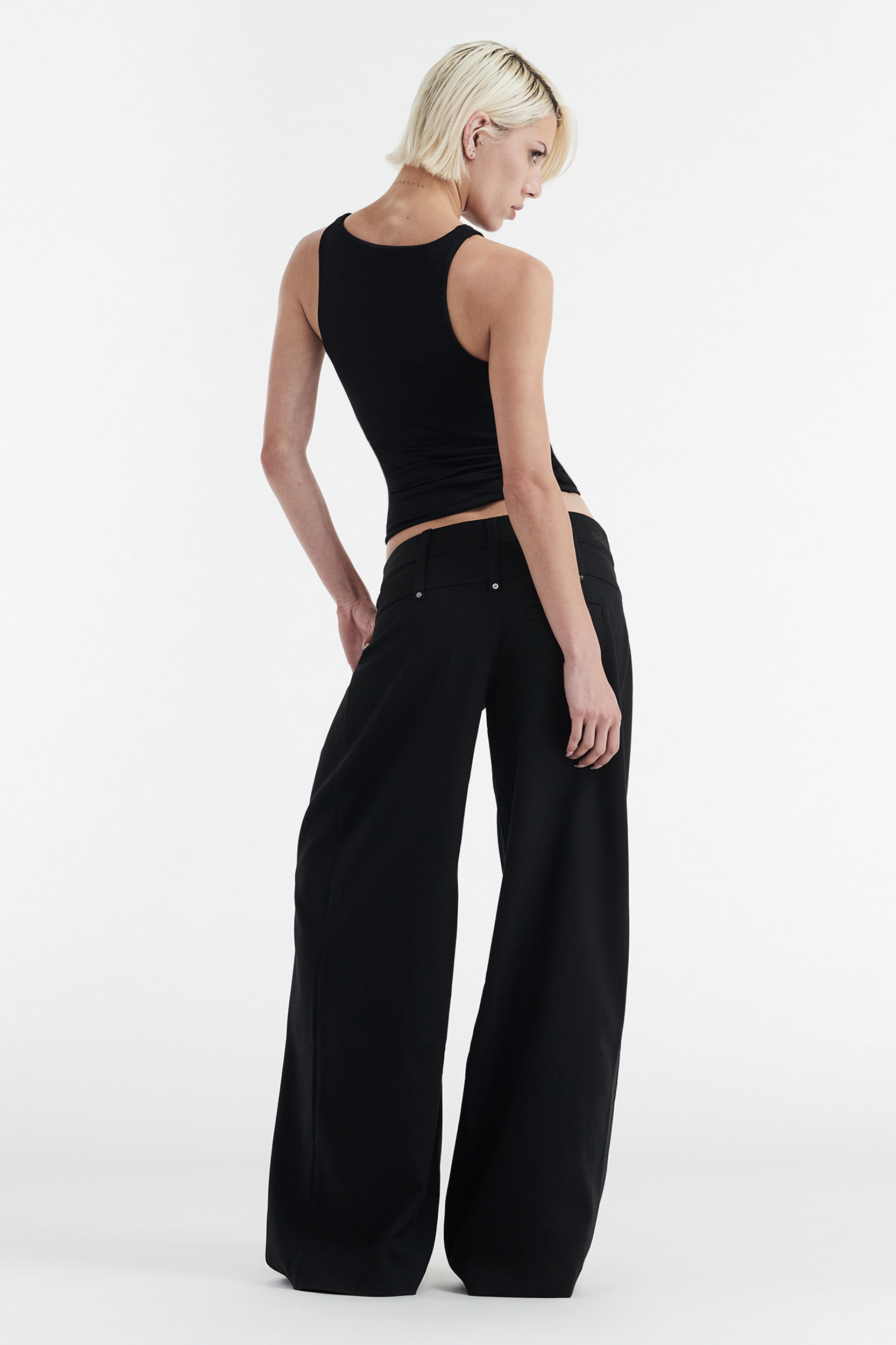 A person with short blonde hair wears the CDP SOPHIE BASIC SLEEVELESS TANK WITH SHELF BRA in black, paired with loose-fitting black pants, standing with their back to the camera against a plain white background.