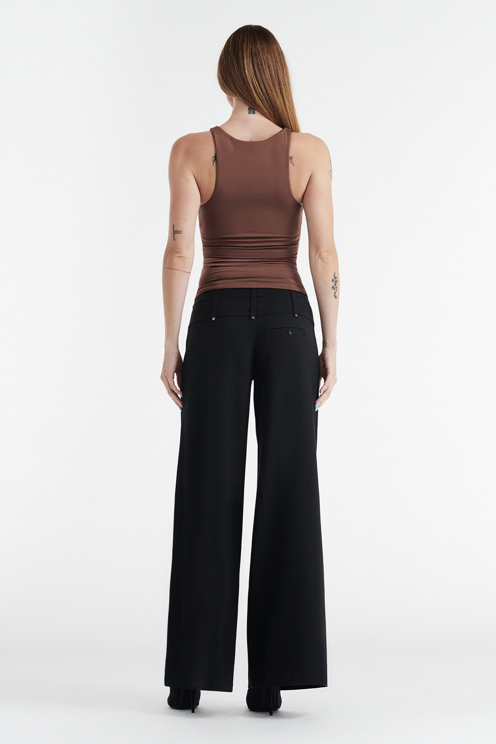 A person stands facing away in the CDP SOPHIE BASIC SLEEVELESS TANK WITH SHELF BRA in chocolate, paired with high-waisted black wide-leg trousers and black shoes, set against a plain white background.