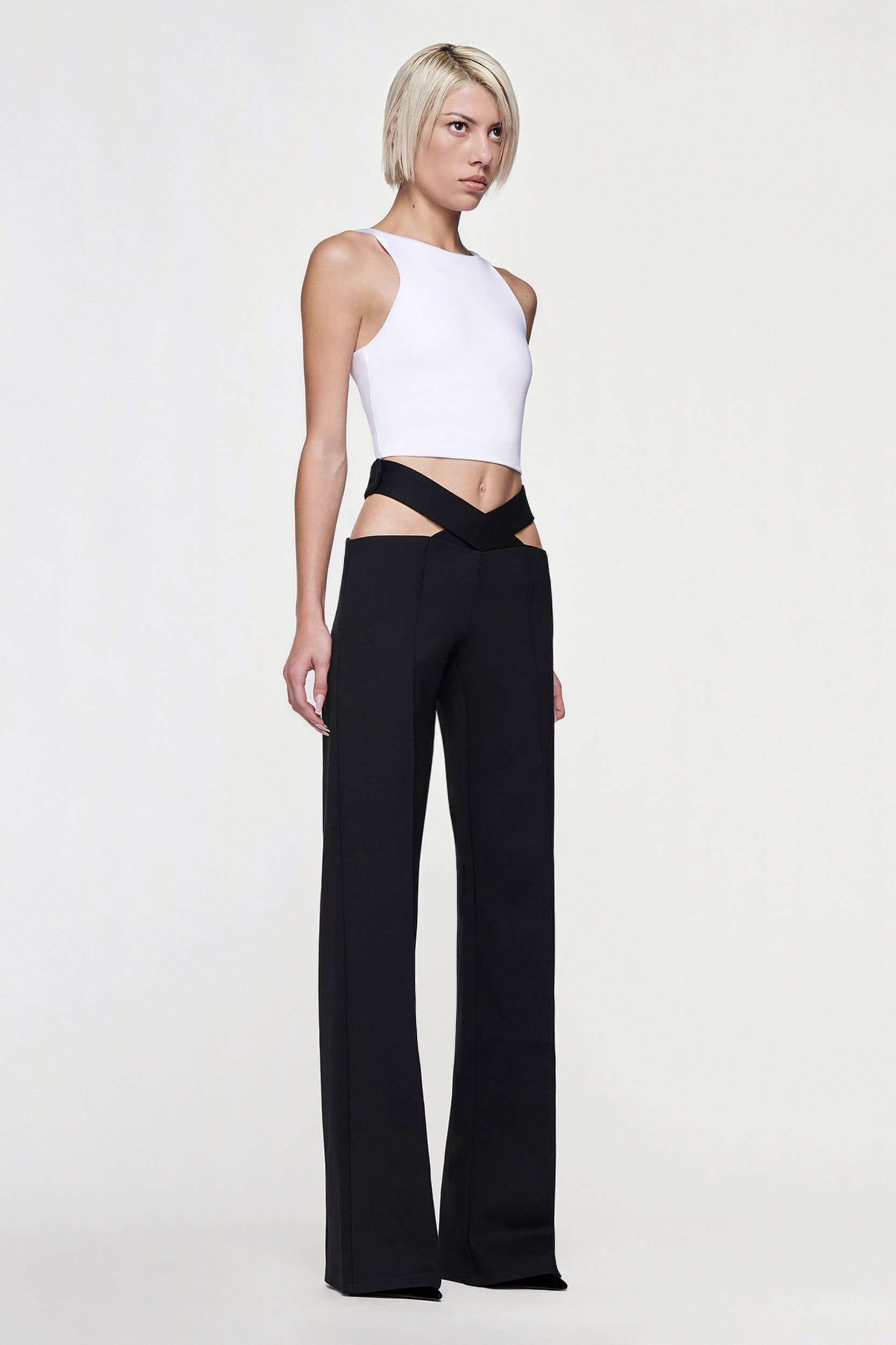A woman with short blonde hair wears a white sleeveless crop top and the CDP JULIETTE WIDE LEG PANT in black, featuring cut-out hip detailing, standing against a plain white background.