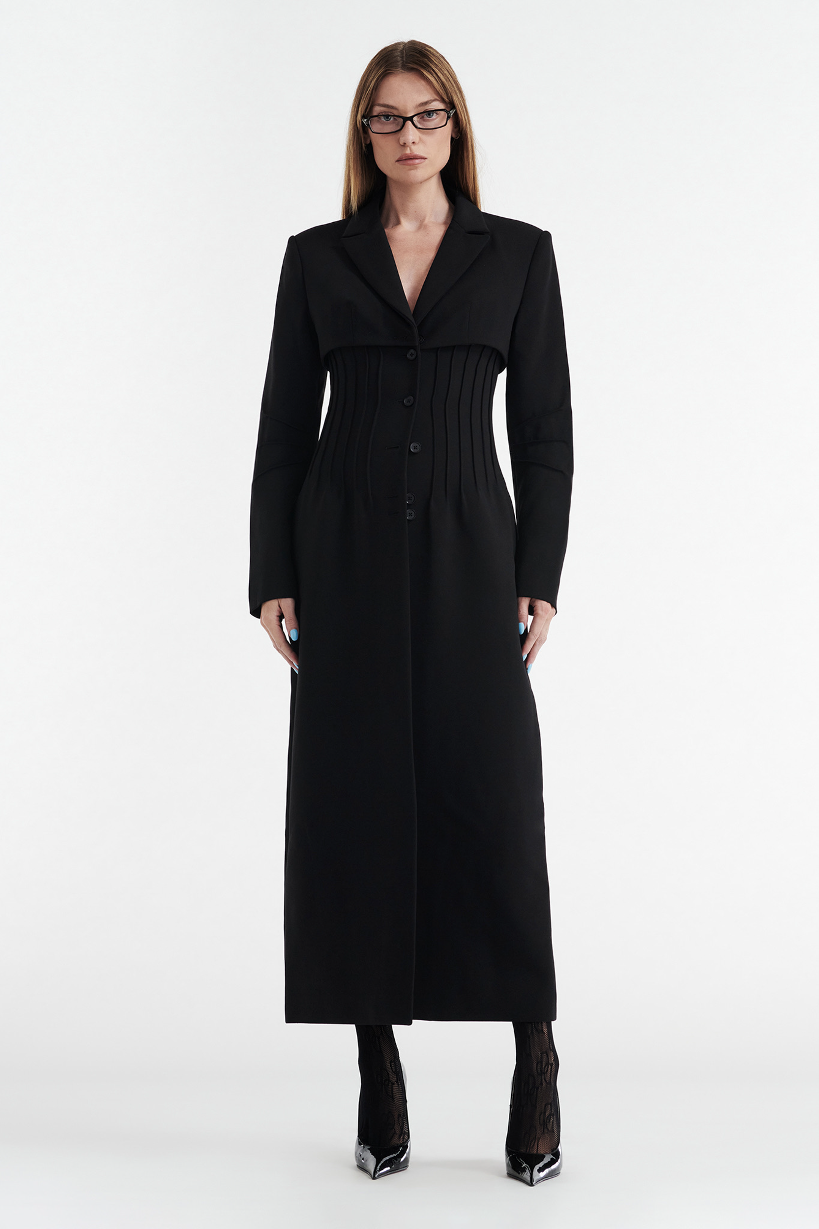 A woman with straight brown hair and glasses wears a CDP CELINE BUTTON UP TRENCH COAT - BLACK, featuring buttons, long sleeves, and a tailored fit. She faces forward in black heels and sheer stockings against a white background.