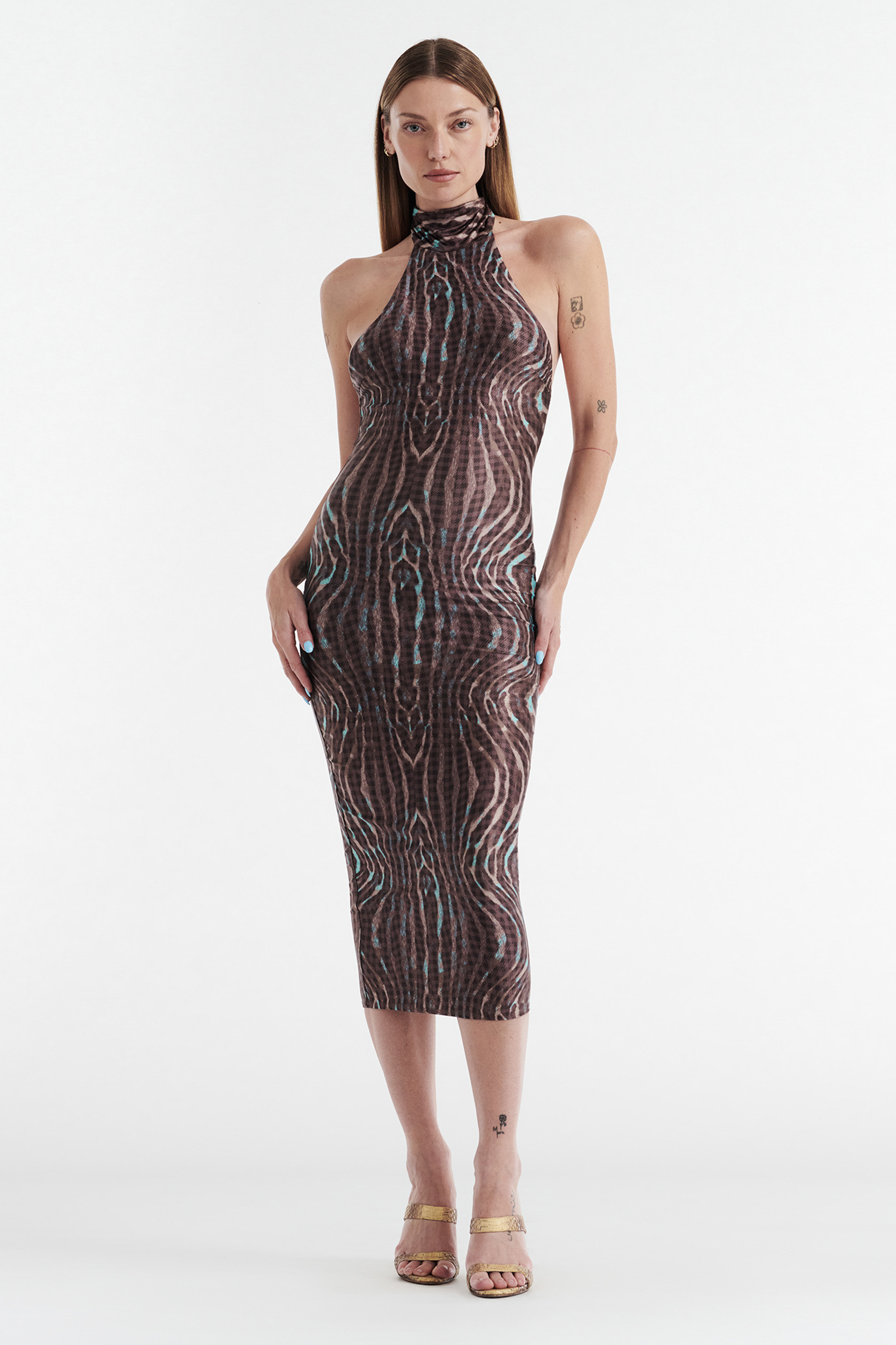 A woman stands against a white background wearing the CDP CLEO Halter Tie Midi Dress in Warped Check, a fitted sleeveless dress with a brown and black geometric pattern. She has straight hair, tattoos, and gold open-toe heels.