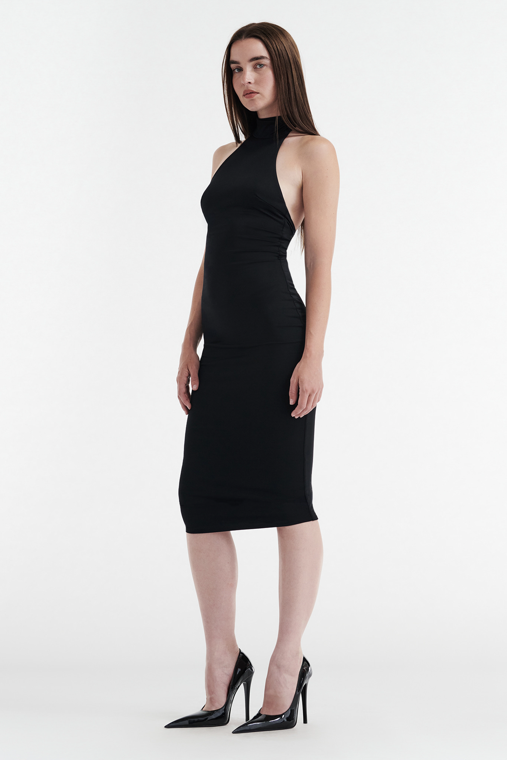 A woman with straight brown hair wears the CDP CLEO Halter Tie Midi Dress in black, paired with black high-heeled shoes, standing against a plain white background.