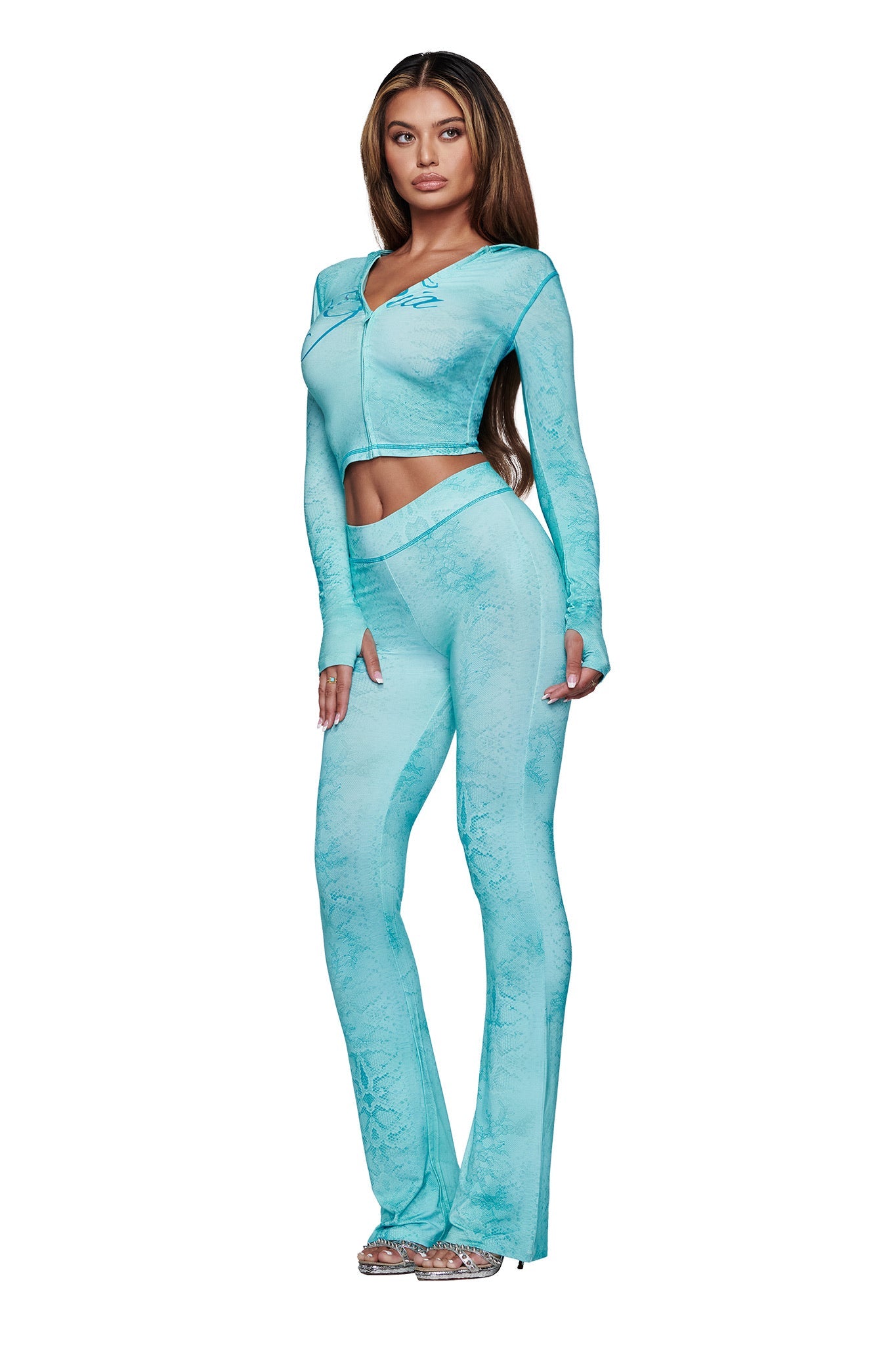 A woman faces forward in the I.AM.GIA BLARE HOODIE - TEAL SERPENTINE, paired with matching high-waisted, flared pants and clear platform heels. She has long wavy hair and a neutral expression.