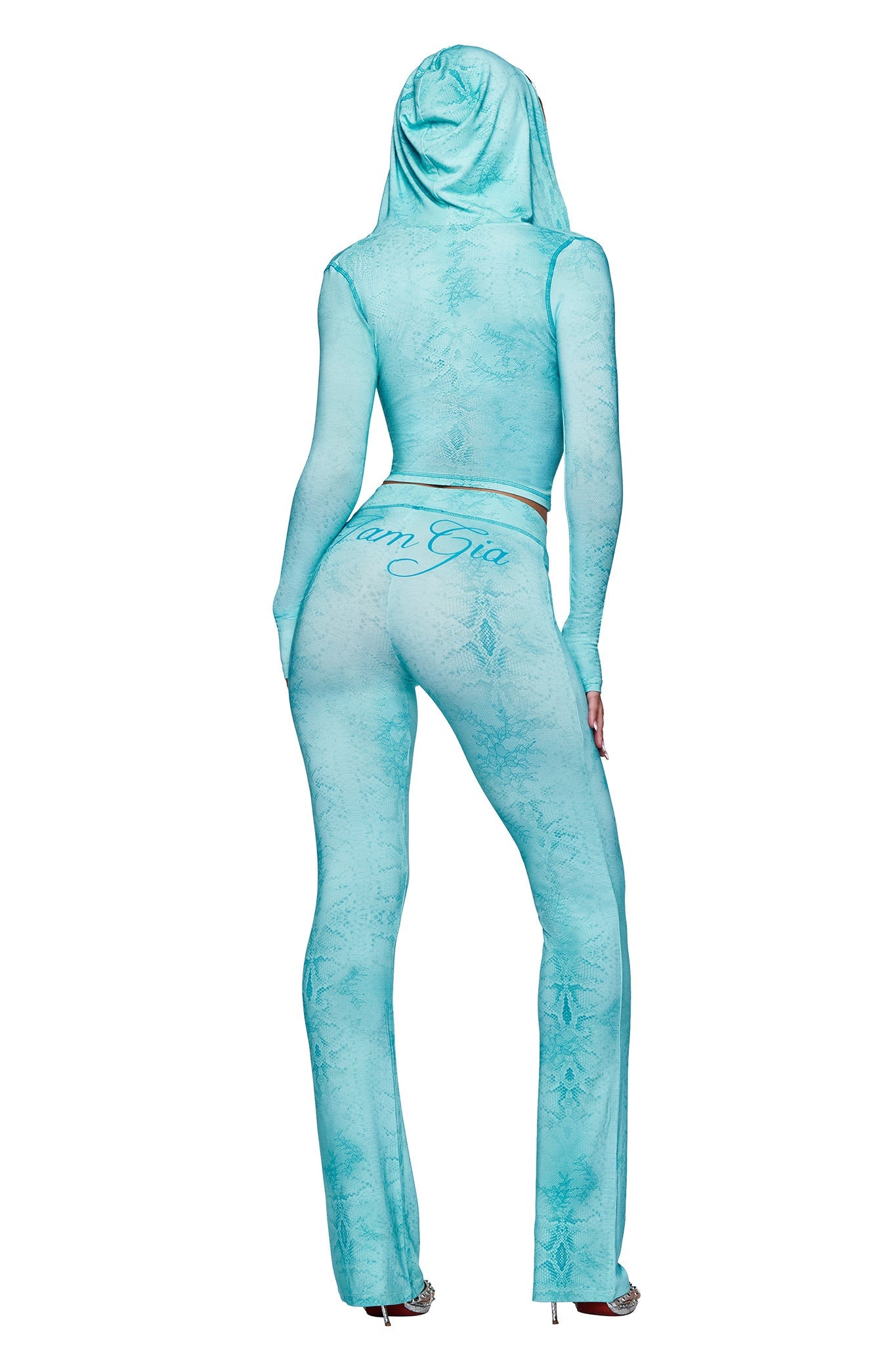 A person stands facing away, wearing fitted, sheer, teal serpentine Blare Trackpants by I.AM.GIA with a script logo across the back. The outfit features long sleeves, flared legs, and is paired with high heels.