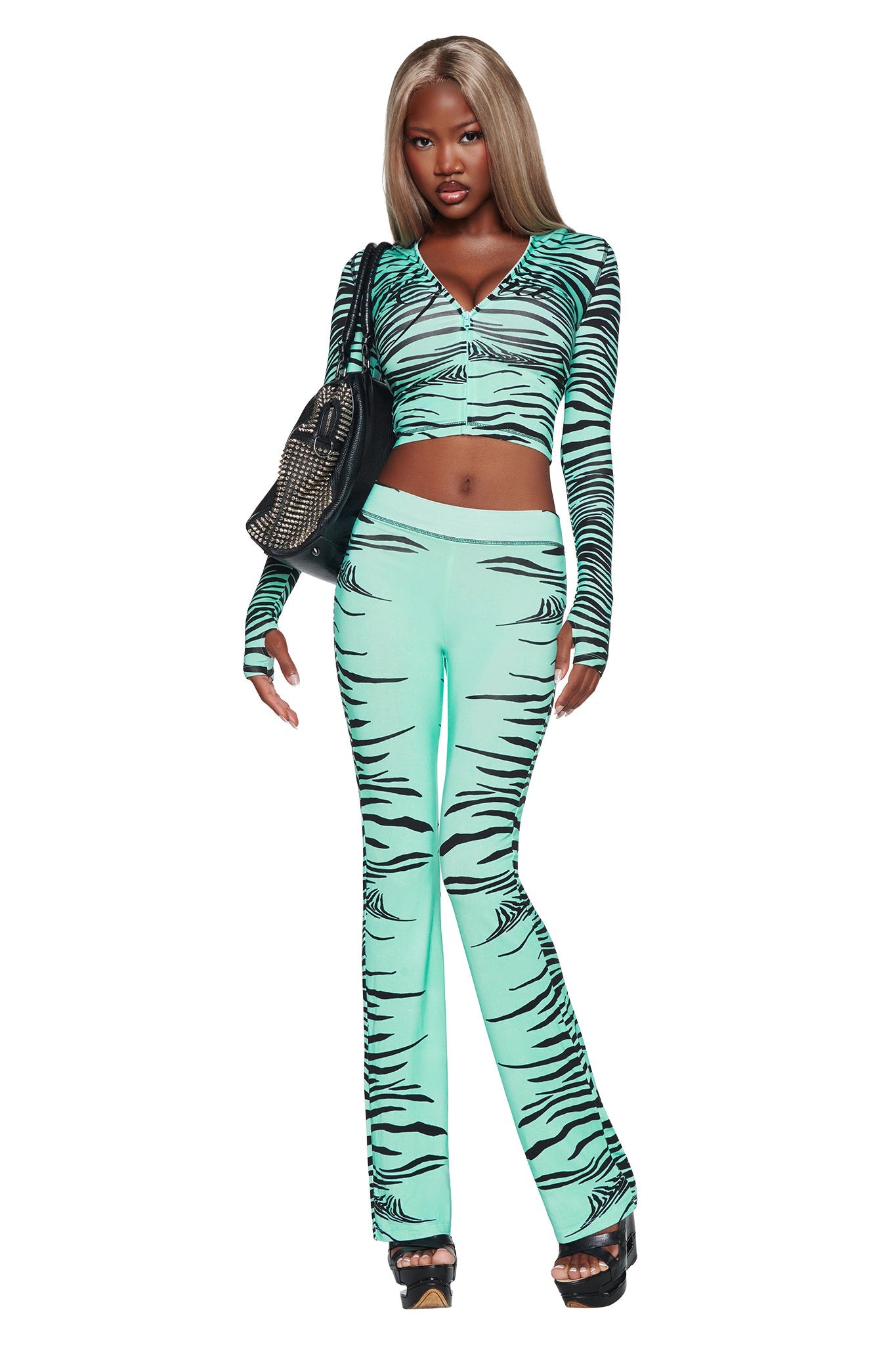 A woman with long blonde hair wears the I.AM.GIA BLARE HOODIE in Aqua Fine Line Zebra—a turquoise and black tiger-striped design—paired with matching pieces, black platform sandals, and a black bag. She stands confidently against a white background.