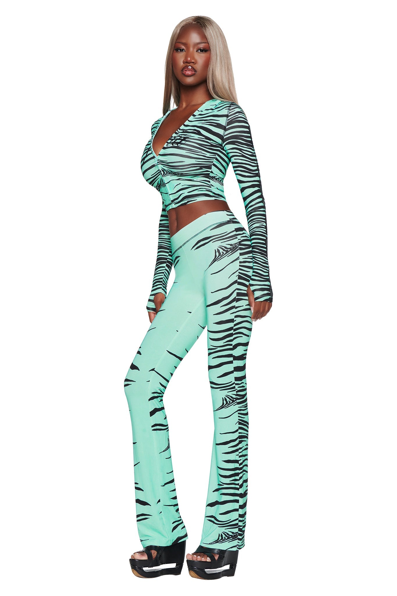A woman with long blonde hair wears the I.AM.GIA BLARE HOODIE in Aqua Fine Line Zebra, featuring a mint green and black print. She pairs it with black platform sandals and stands confidently, facing slightly to the side.