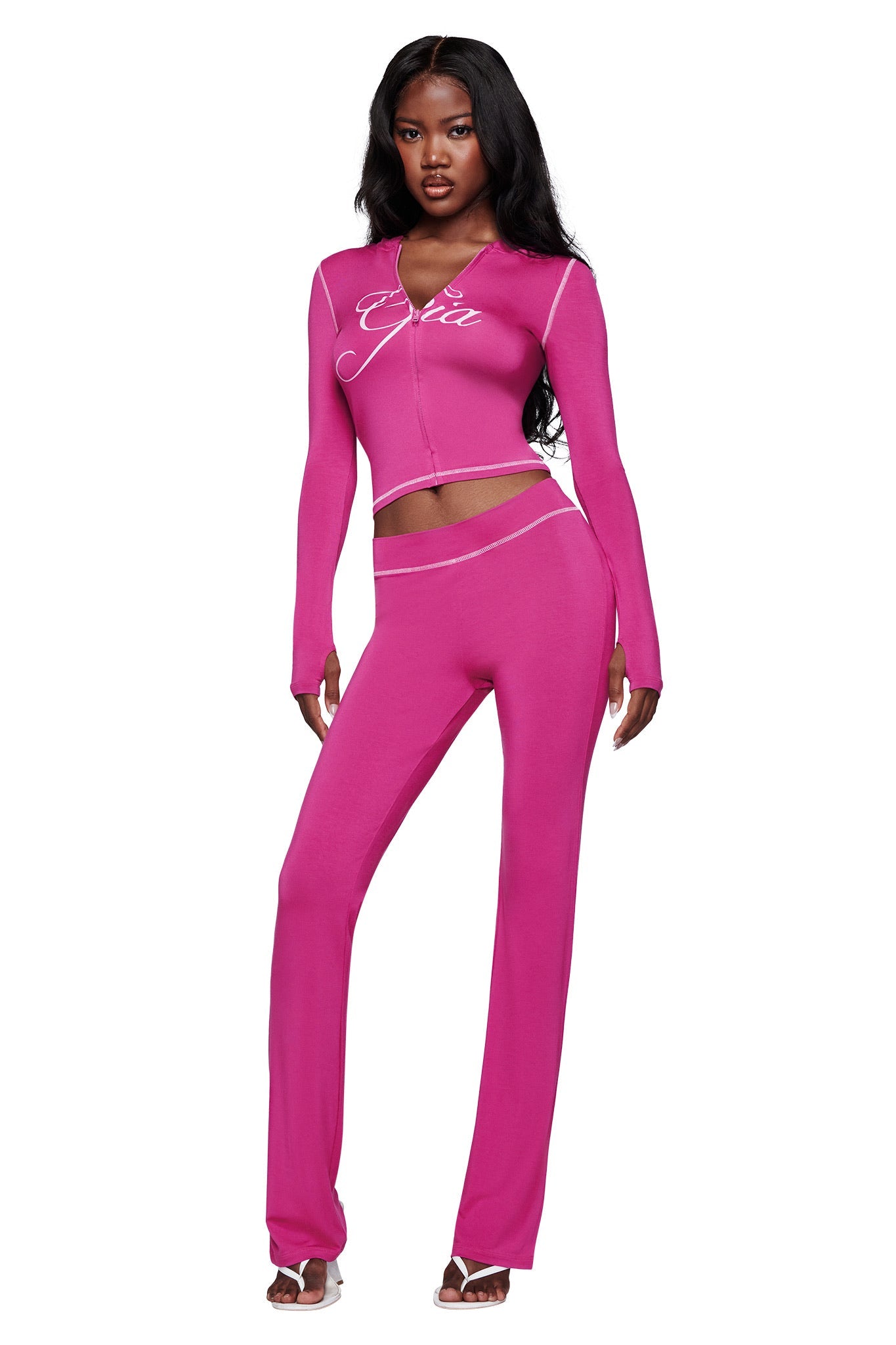 A woman wears the I.AM.GIA BLARE HOODIE in Raspberry, a bright pink zip-up with white script text and piping, paired with matching flared pants, white heeled sandals, and long wavy hair.