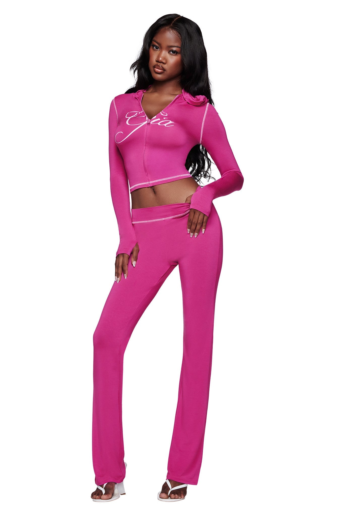 A woman poses in the I.AM.GIA BLARE HOODIE in raspberry with matching pants and white heeled sandals, her long wavy dark hair flowing against a plain white background.
