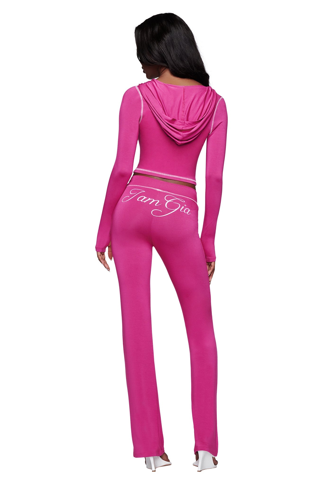 A woman is seen from the back wearing the I.AM.GIA BLARE HOODIE in Raspberry—a bright pink, fitted hoodie tracksuit. The matching pants feature "I Am Gia" in white cursive on the waistband, styled with white heeled shoes for a bold look.