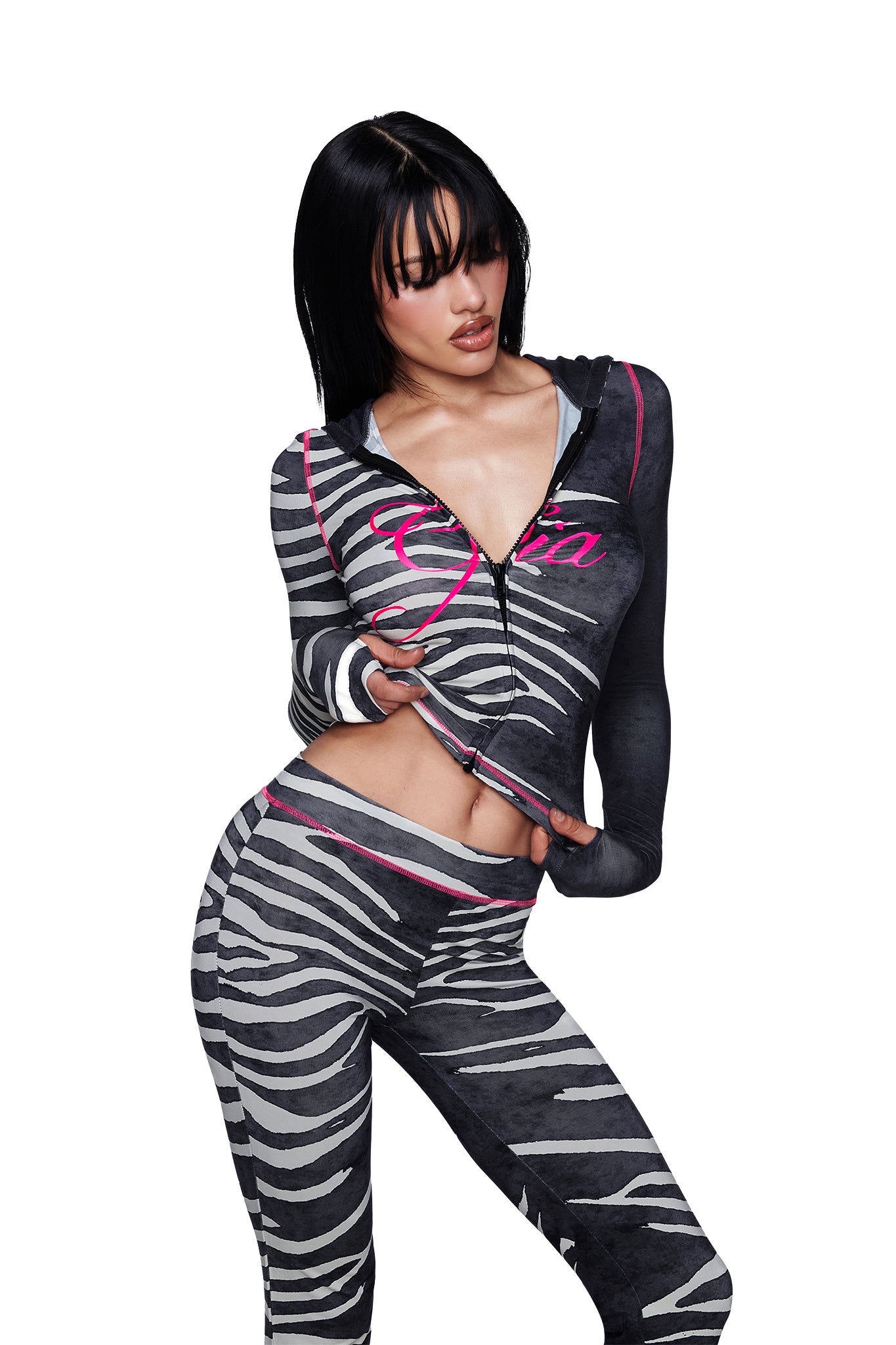 A woman poses in a zebra-print outfit featuring the I.AM.GIA BLARE TRACKPANT - BLACK WILD SIDE paired with a zip-up hoodie, exposing part of her midriff. She has dark hair and is looking downward with her mouth slightly open.
